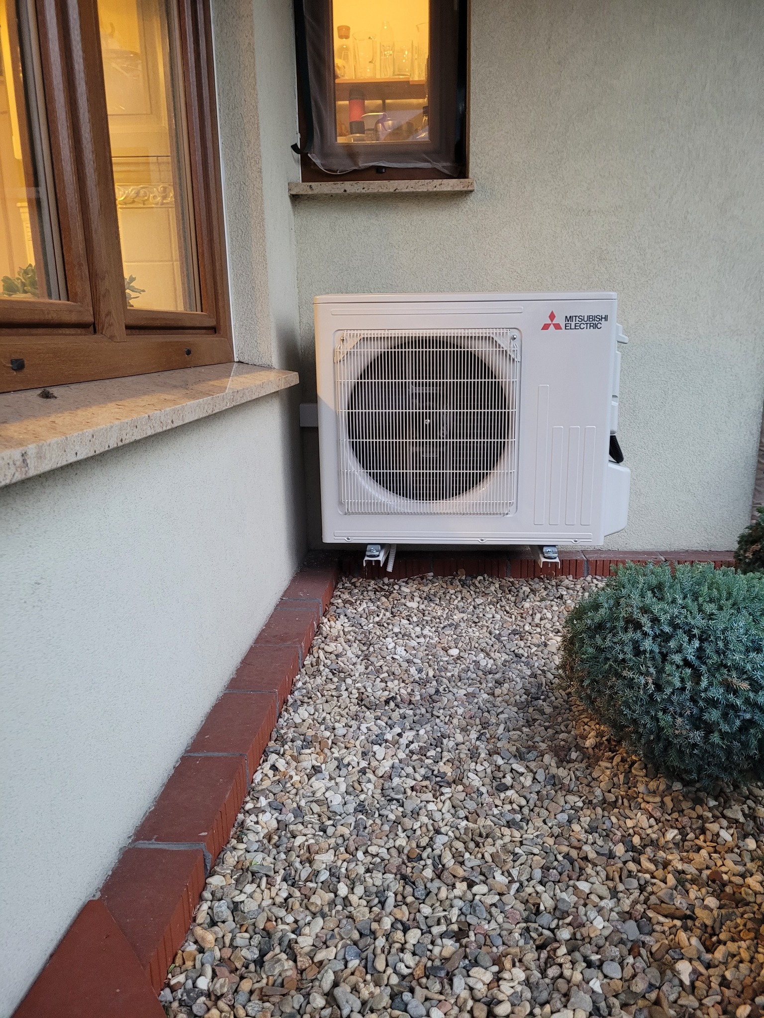 Zewnętrzna jednostka klimatyzacji Mitsubishi Electric zamontowana na ścianie budynku, otoczona ozdobnym żwirem i niskim krzewem.