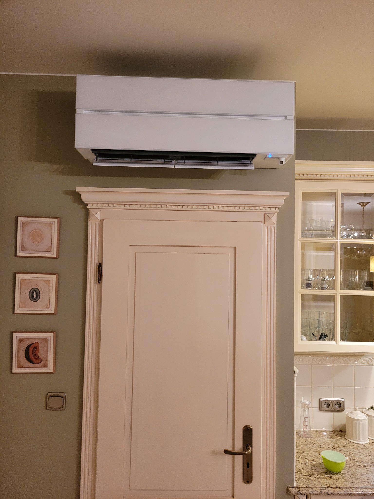 Biały klimatyzator ścienny zamontowany nad kremowymi drzwiami w pomieszczeniu z elementami klasycznej zabudowy kuchennej.