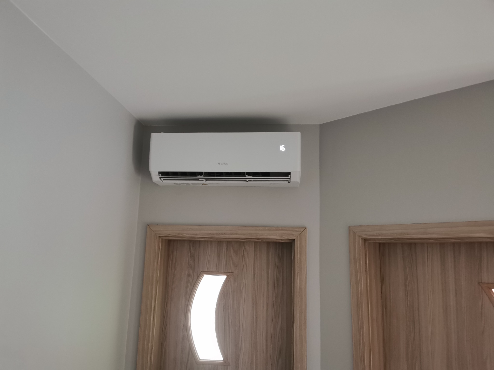 Biały klimatyzator marki Gree zamontowany na ścianie w pokoju z dwiema drewnianymi ościeżnicami drzwi, wyświetlający temperaturę 16 stopni Celsjusza.