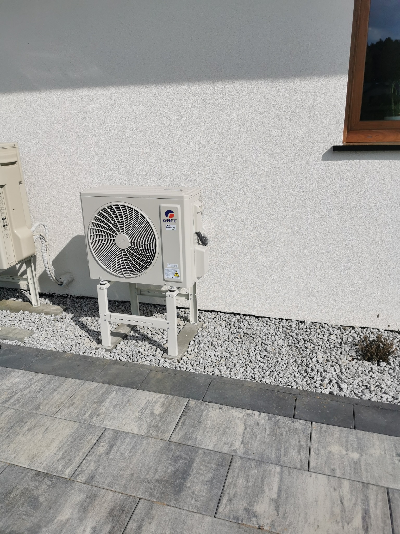 Zewnętrzna jednostka klimatyzacji marki Gree zamontowana na białym cokole przy białej ścianie domu, otoczona drobnym białym żwirem i szarym chodnikiem z kostki. Widoczna również druga jednostka...