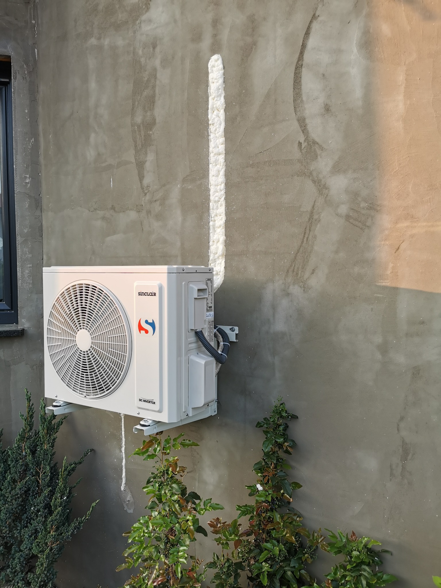 Zewnętrzna jednostka klimatyzacji Sinclair zamontowana na surowej, szarej ścianie budynku z widoczną pianką montażową uszczelniającą przewody, z roślinami ozdobnymi u podstawy ściany.