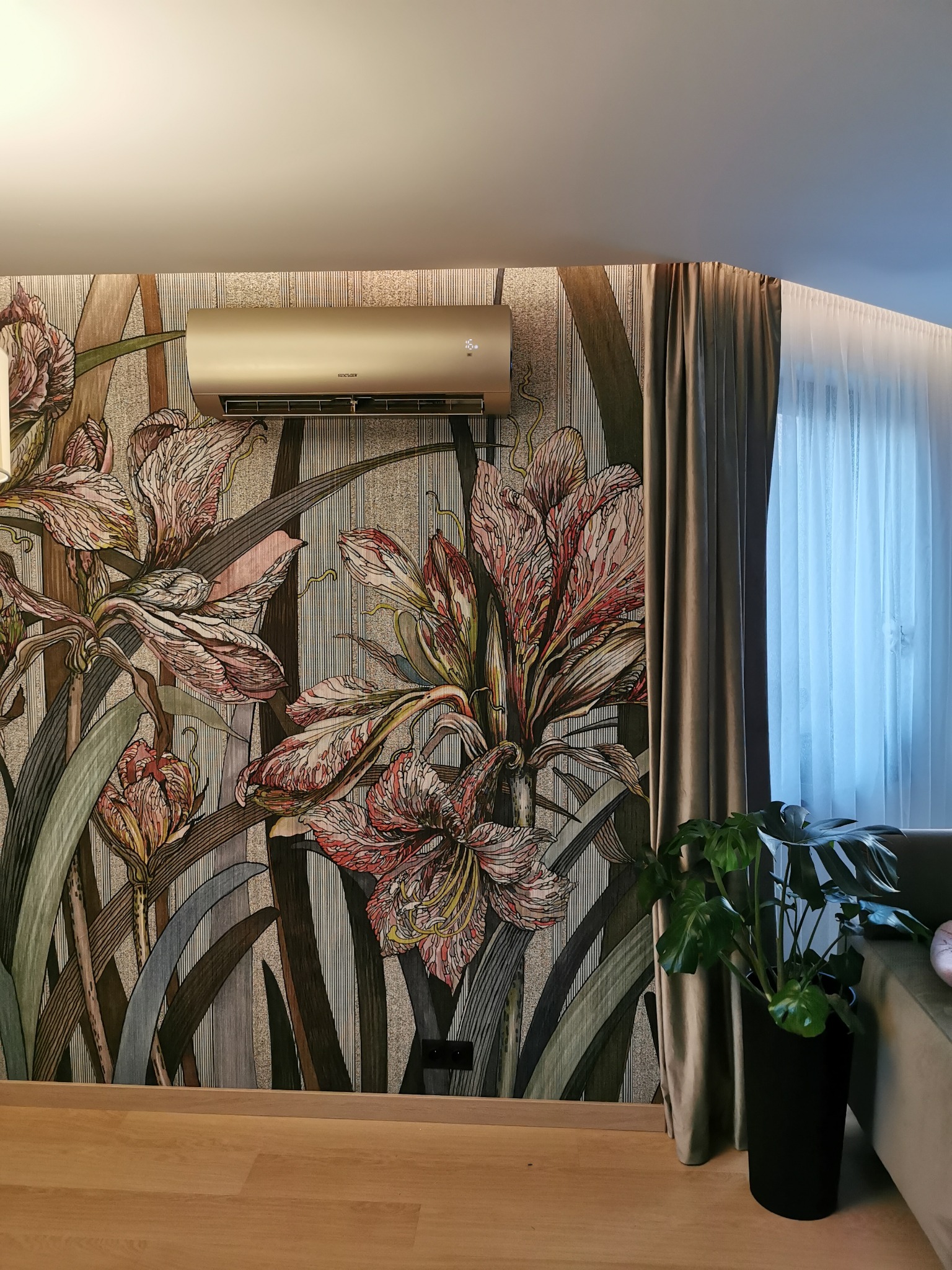 Złoty klimatyzator Sinclair zamontowany na ścianie z wzorem kwiatowym, obok zasłony i rośliny doniczkowej w salonie.