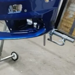 Błyszczący, niebieski zderzak samochodowy na wózku warsztatowym, po lakierowaniu, widoczne mocowania i czujnik parkowania.