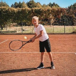 Marta Ziętek Fotografia - Uśmiechnięty chłopiec w białej koszulce i czarnych spodenkach odbija piłkę tenisową rakietą na korcie ziemnym w słoneczny dzień.
