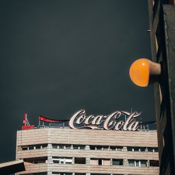Marta Ziętek Fotografia - Budynek z neonowym logo Coca-Cola na dachu, widok z dołu, lampa uliczna w prawym rogu kadru, pochmurne niebo w tle.