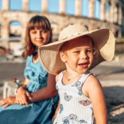Dwie dziewczynki siedzą przed antycznym amfiteatrem w Puli, Chorwacja, młodsza dziewczynka w słomkowym kapeluszu i sukience w motyle uśmiecha się do kamery.