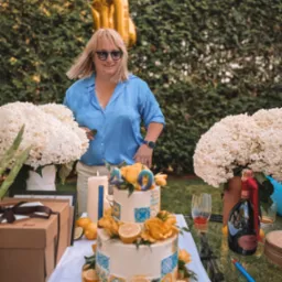 Uśmiechnięta kobieta w okularach przeciwsłonecznych i błękitnej koszuli stoi za stołem z tortem urodzinowym, kwiatami hortensji, prezentami i butelką szampana Ruinart, na tle zielonego żywopłotu.