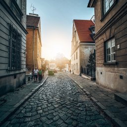 Marta Ziętek Fotografia - Ulica z brukowaną nawierzchnią w starej części miasta, z budynkami po obu stronach i grupą ludzi spacerujących w oddali w kierunku słońca.