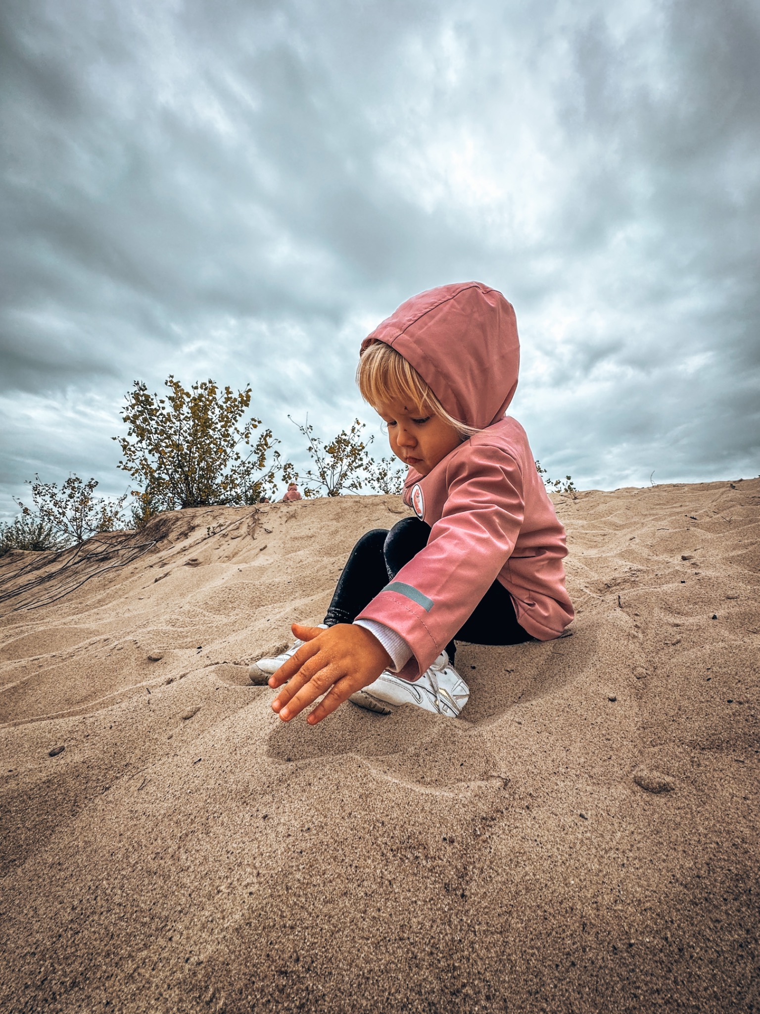 Dziewczynka w różowej kurtce z kapturem siedzi na piaszczystej wydmie, bawiąc się piaskiem na tle pochmurnego nieba.