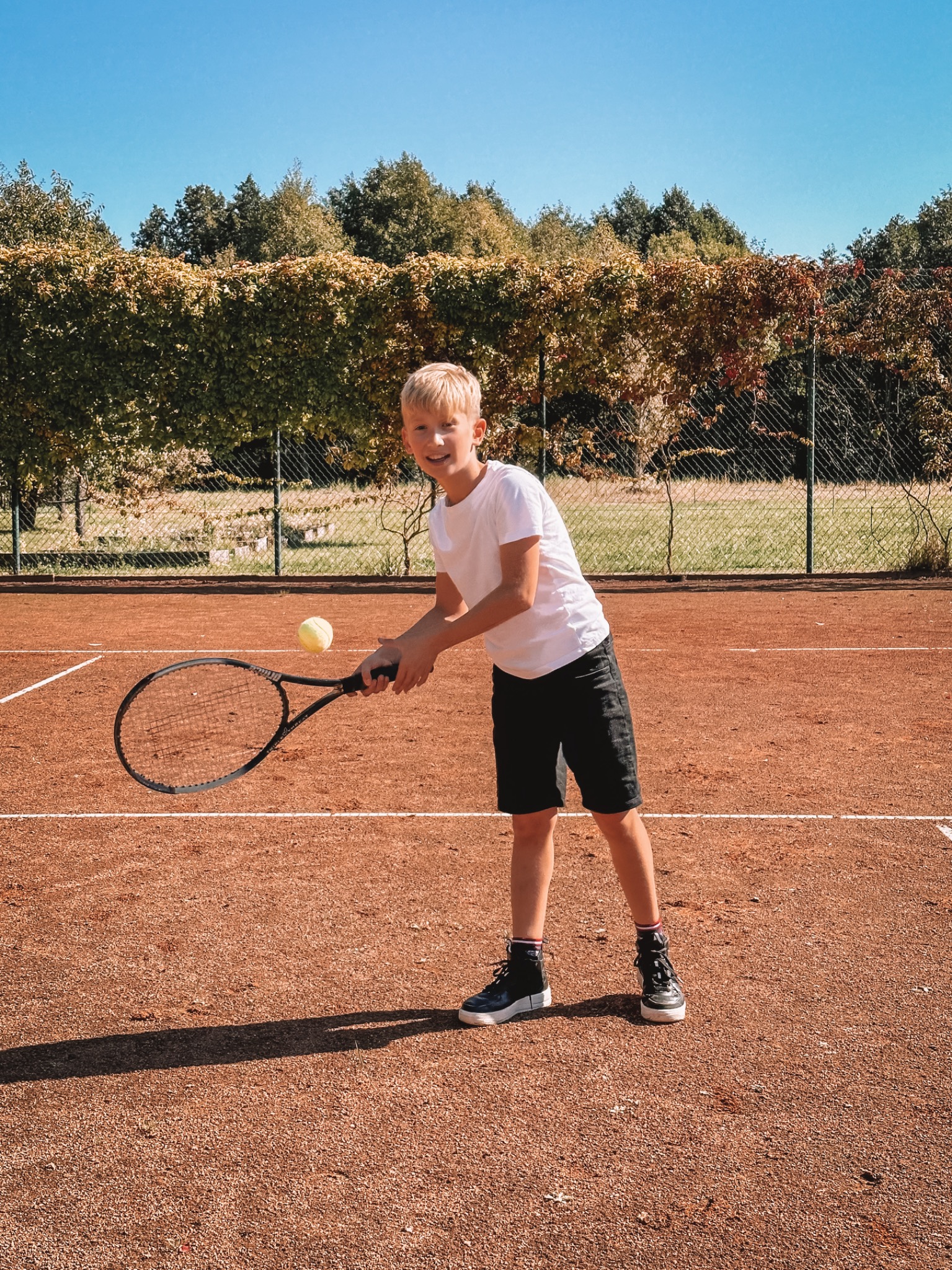 Uśmiechnięty chłopiec w białej koszulce i czarnych spodenkach odbija piłkę tenisową rakietą na korcie ziemnym w słoneczny dzień.
