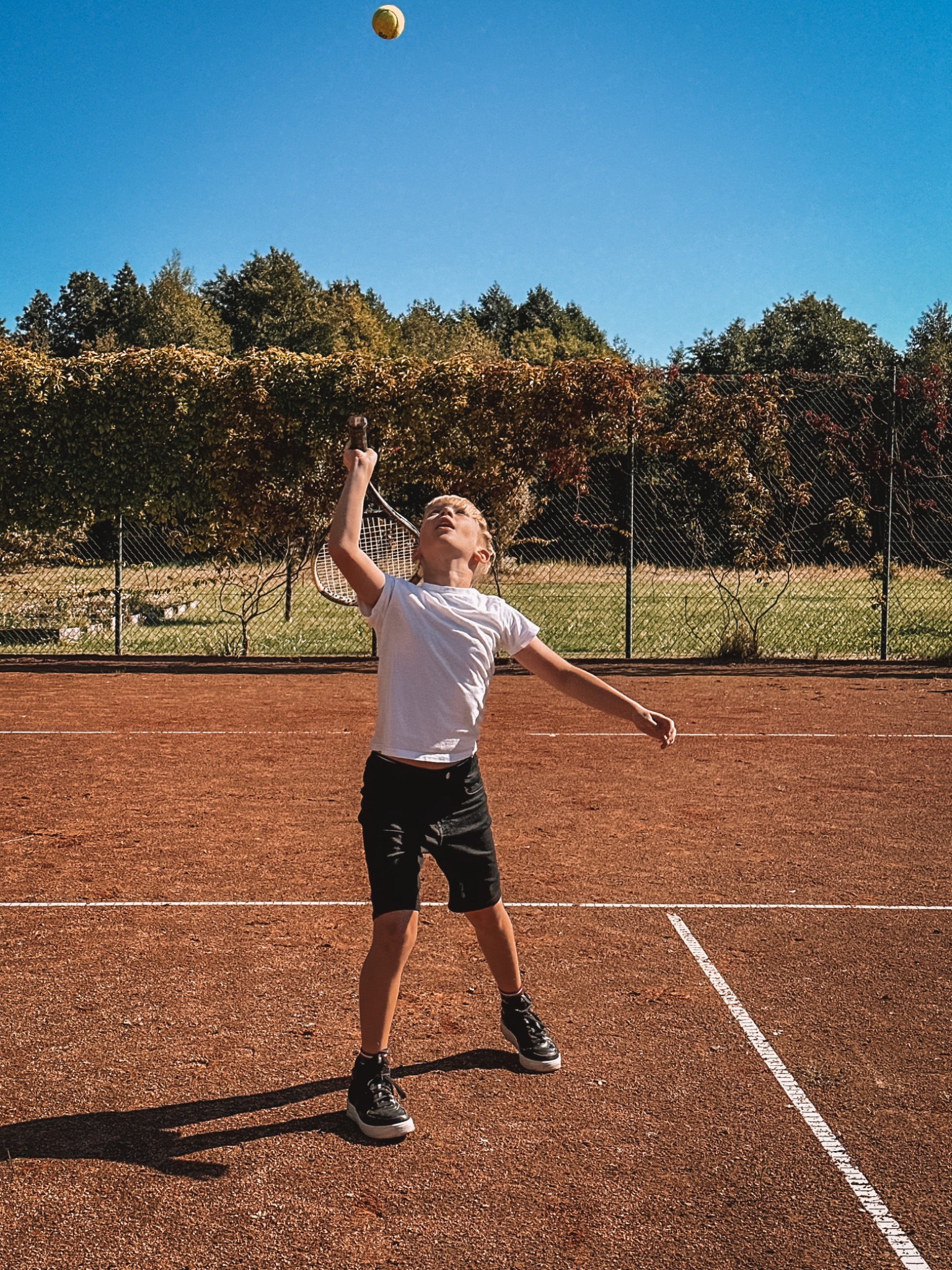 Chłopiec w białej koszulce i czarnych spodenkach uderza rakietą tenisową w żółtą piłkę na korcie ziemnym w słoneczny dzień.