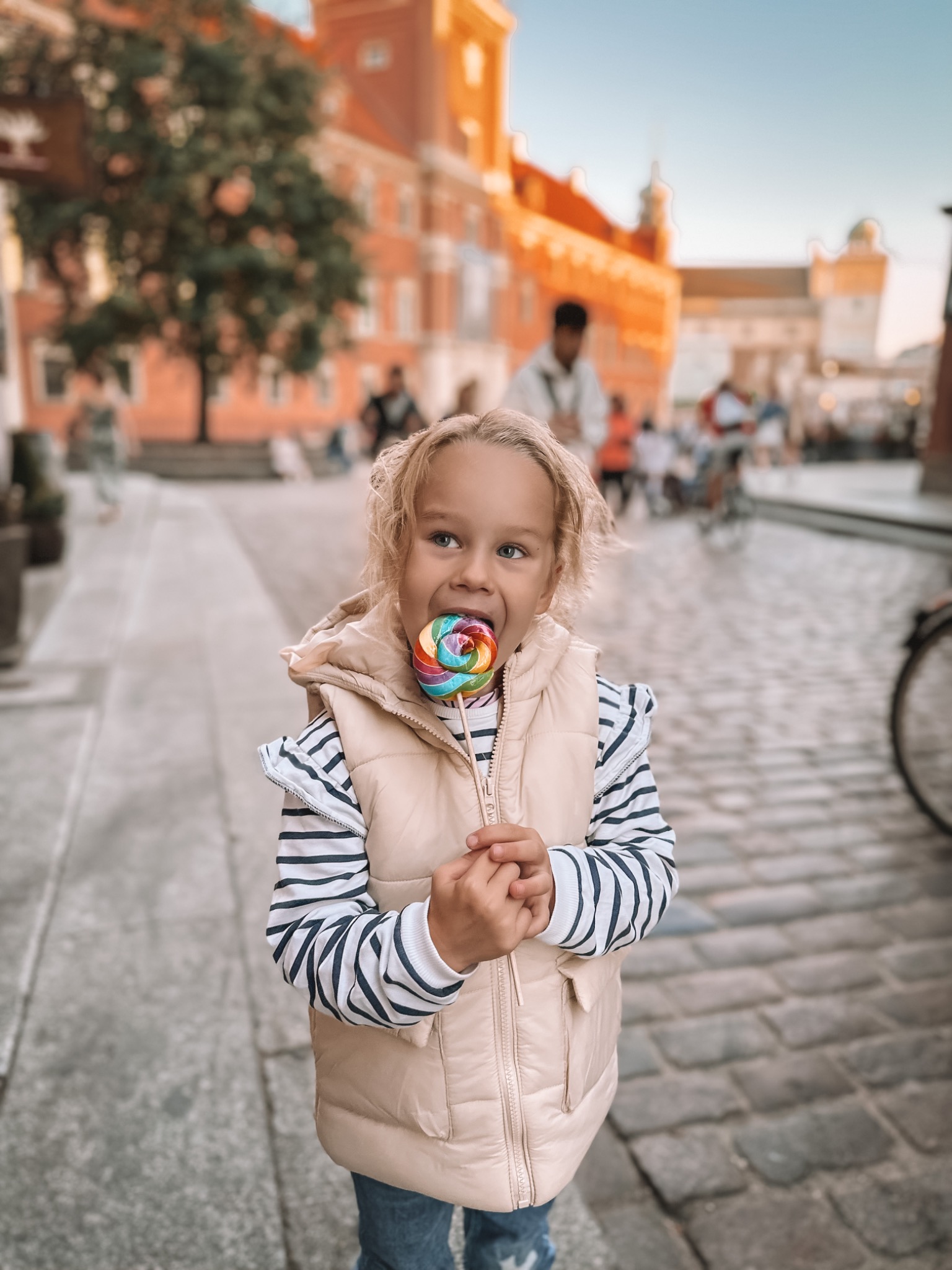 Uśmiechnięta blondwłosa dziewczynka z lizakiem w ustach, ubrana w beżową kamizelkę i bluzkę w paski, na tle brukowanej ulicy i ceglanych budynków w słoneczny dzień.