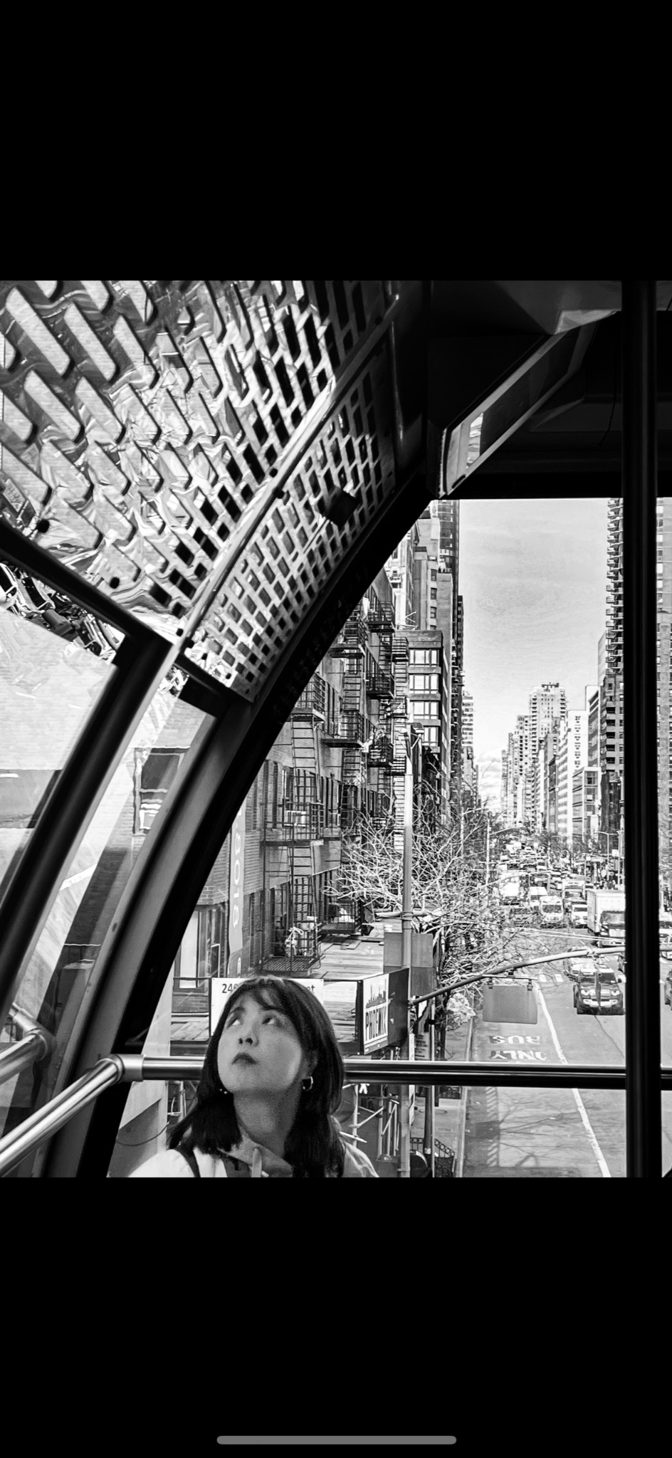 Czarno-białe zdjęcie kobiety patrzącej w górę z wnętrza pojazdu, z widokiem na ulicę miejską z wysokimi budynkami i ruchem ulicznym.