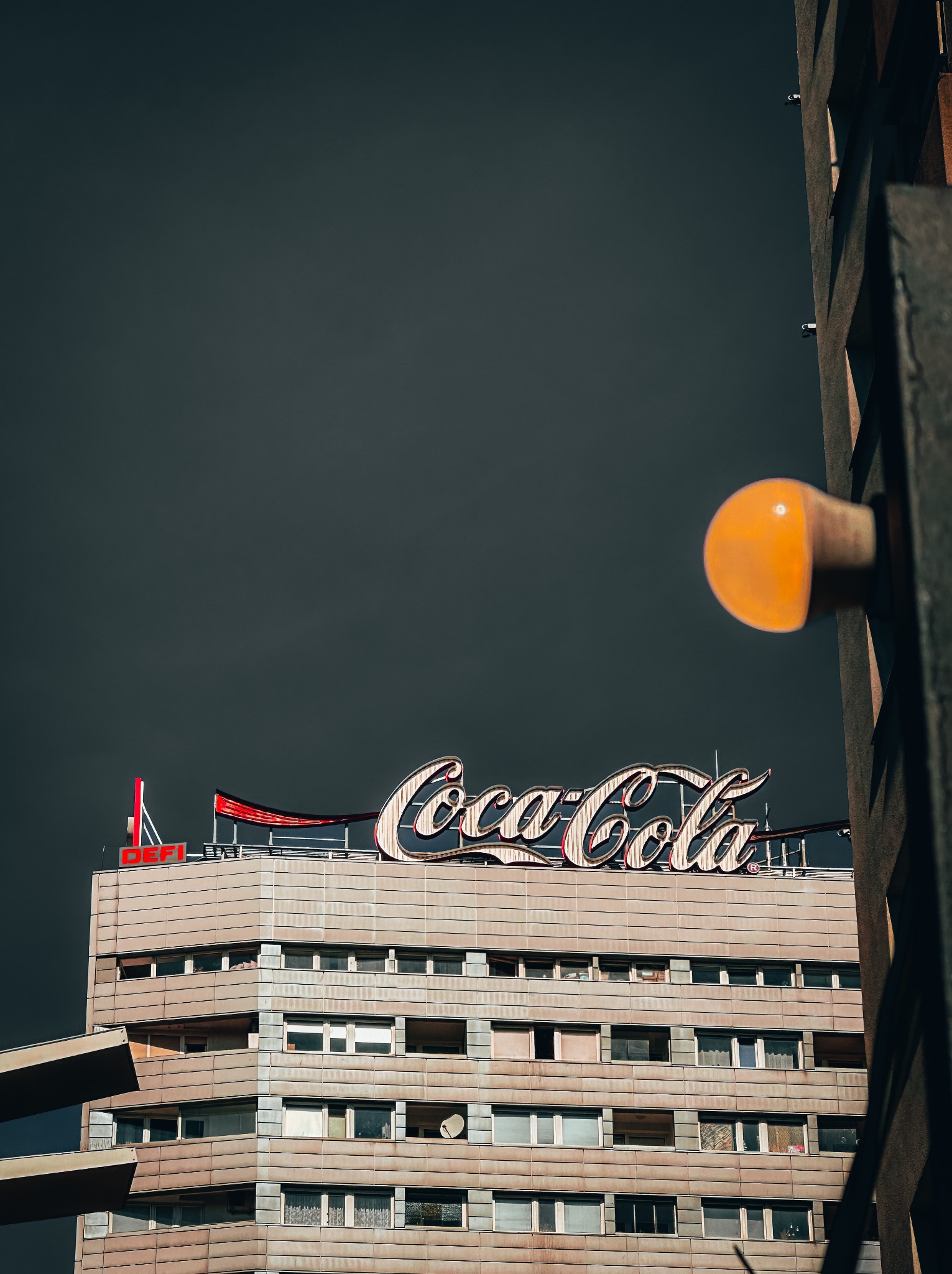Budynek z neonowym logo Coca-Cola na dachu, widok z dołu, lampa uliczna w prawym rogu kadru, pochmurne niebo w tle.