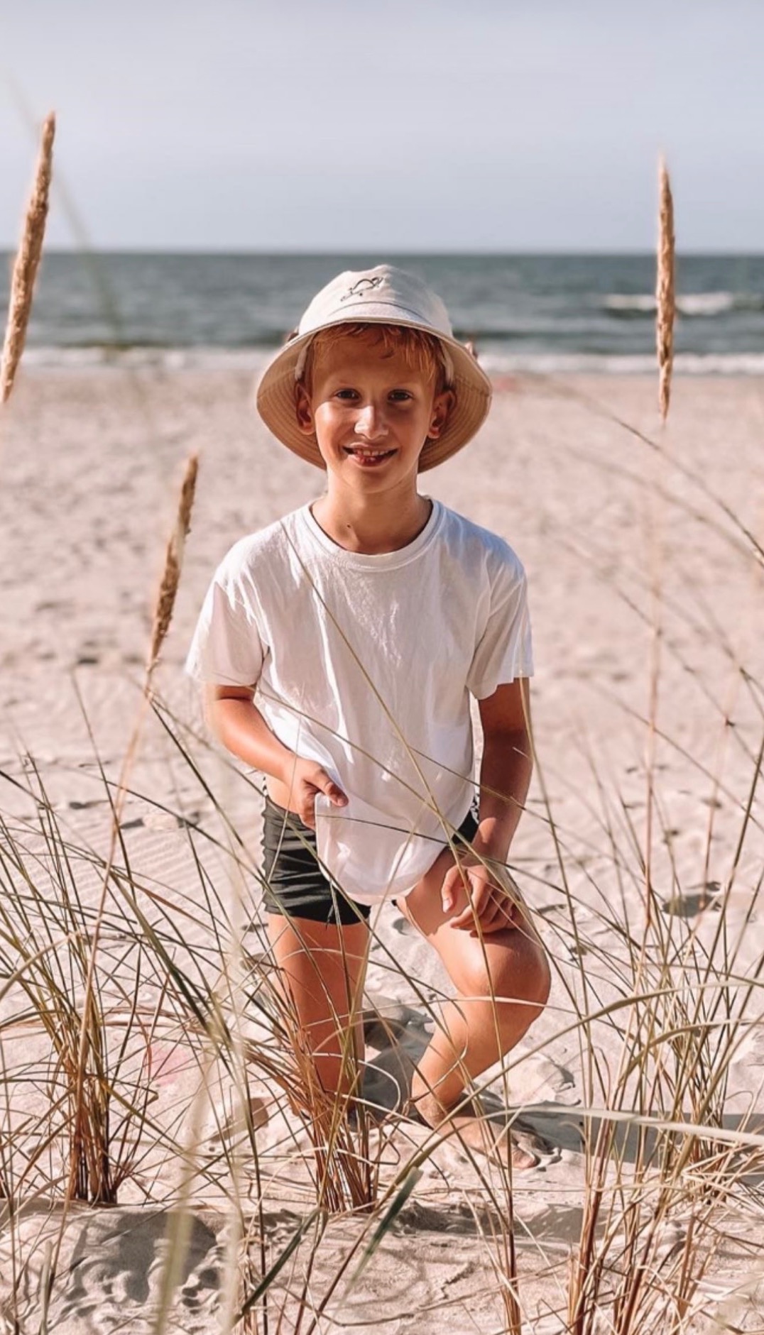 Uśmiechnięty chłopiec w kapeluszu i białej koszulce stoi na piaszczystej plaży, otoczony trawą morską, z widokiem na morze w tle.