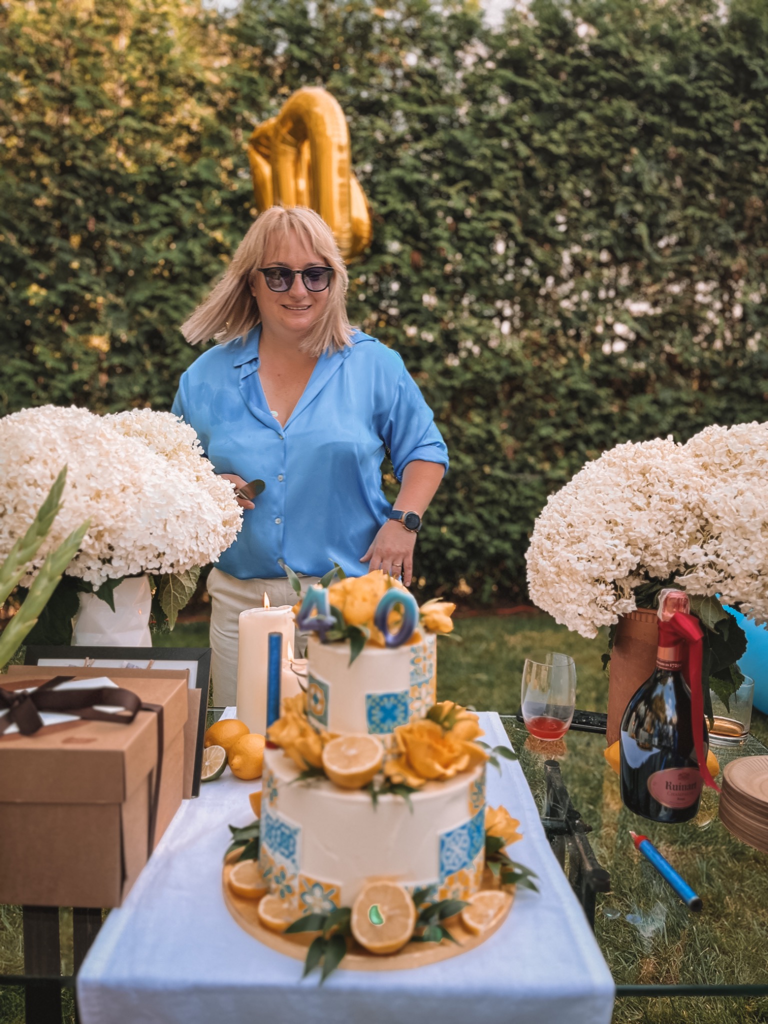 Uśmiechnięta kobieta w okularach przeciwsłonecznych i błękitnej koszuli stoi za stołem z tortem urodzinowym, kwiatami hortensji, prezentami i butelką szampana Ruinart, na tle zielonego żywopłotu.