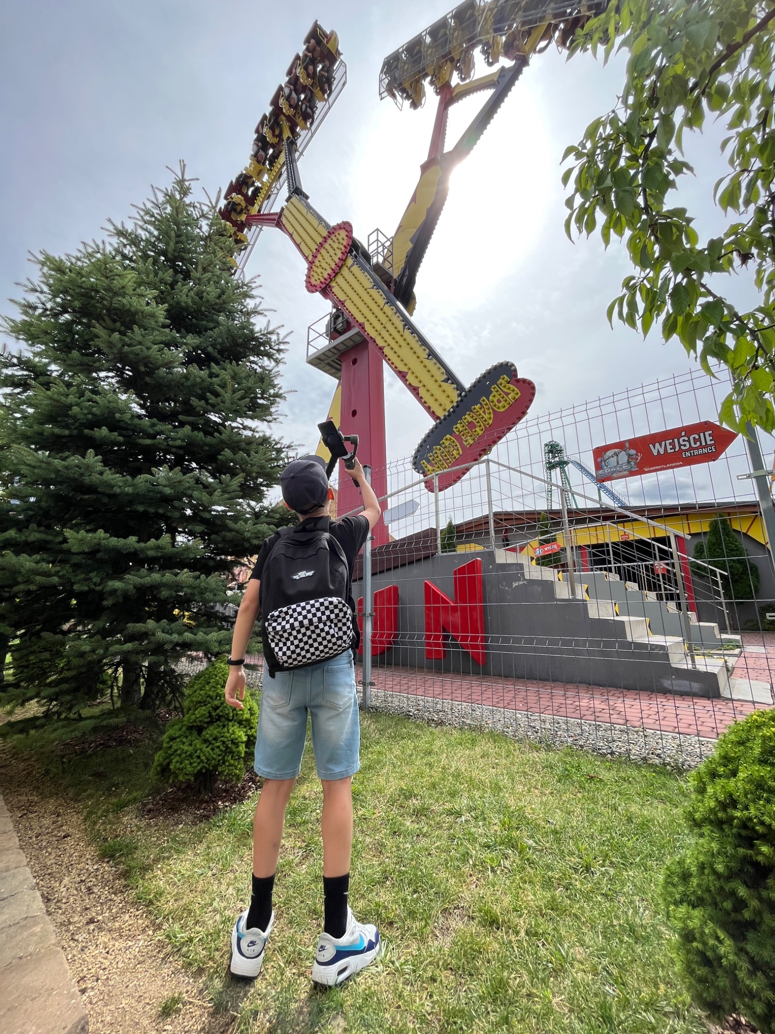 Chłopiec w dżinsowych szortach i czarnej koszulce filmuje telefonem w parku rozrywki dużą karuzelę w kształcie wahadła z ludźmi na pokładzie, na tle zieleni i nieba.