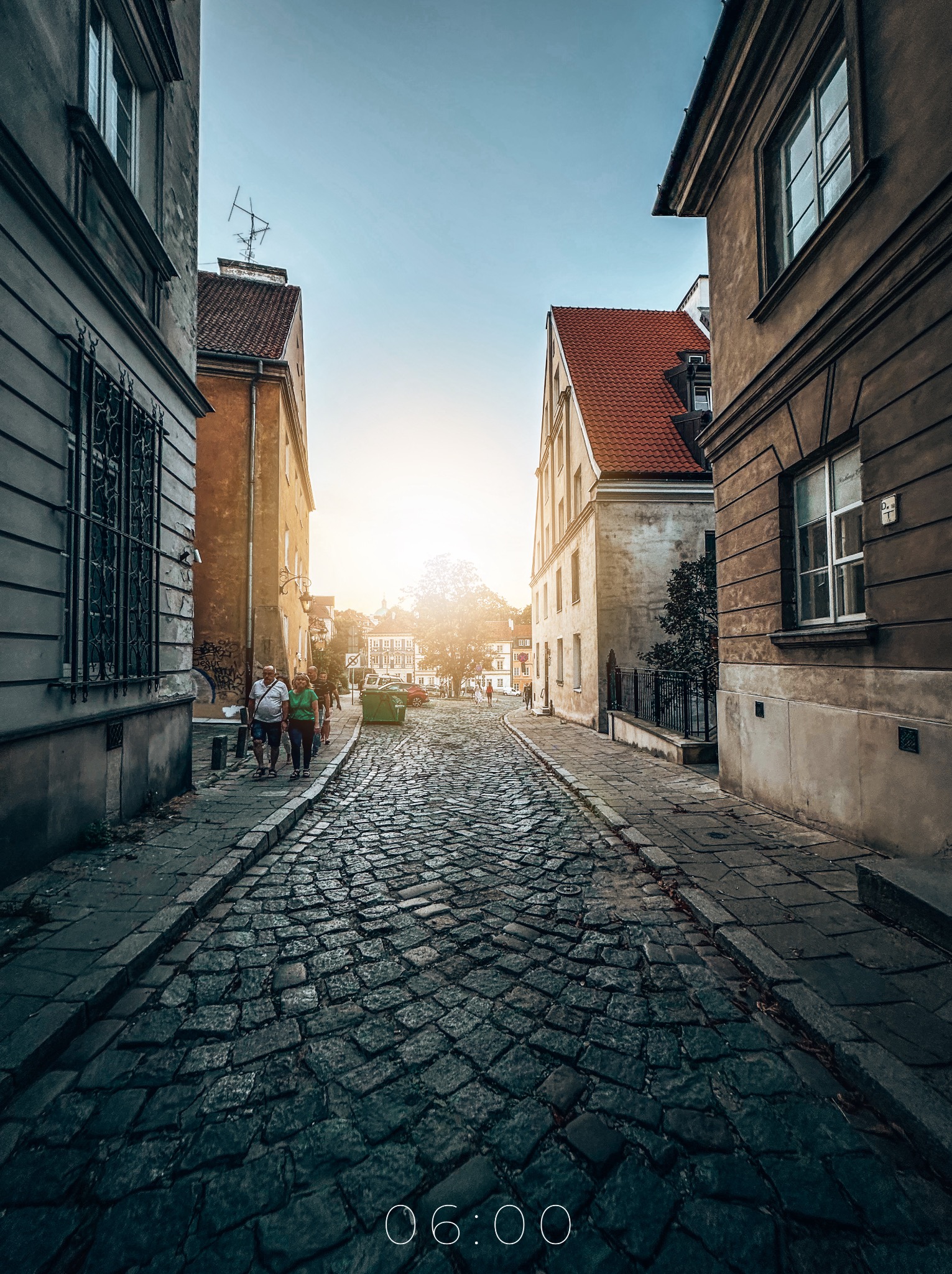 Ulica z brukowaną nawierzchnią w starej części miasta, z budynkami po obu stronach i grupą ludzi spacerujących w oddali w kierunku słońca.