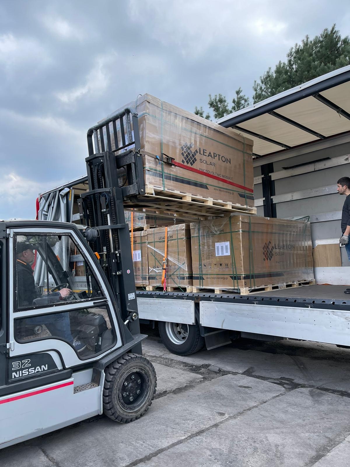 Rozładunek palet z modułami fotowoltaicznymi Leapton Solar z ciężarówki za pomocą wózka widłowego Nissan. Widoczny pracownik nadzorujący proces.