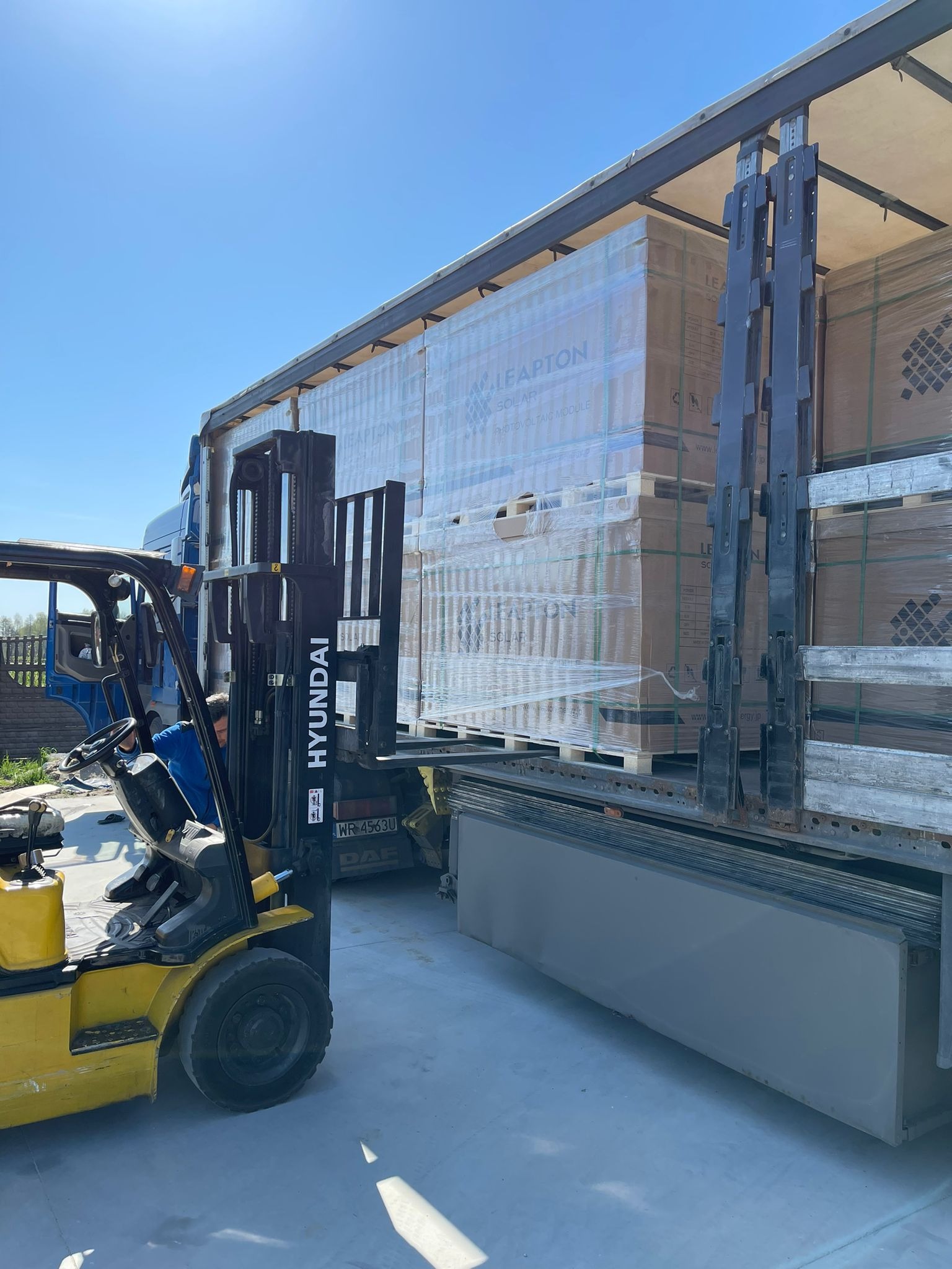 Rozładunek palet z modułami fotowoltaicznymi Leapton z naczepy ciężarówki za pomocą wózka widłowego Hyundai, widoczny operator w niebieskiej koszuli.