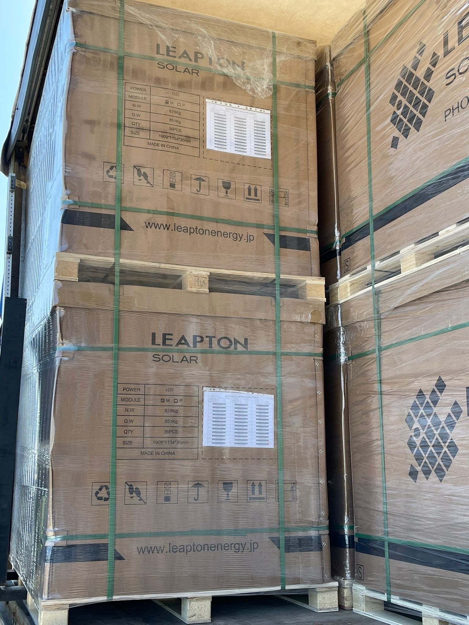 Palety z zapakowanymi modułami solarnymi Leapton Solar, owinięte folią i spięte taśmami, z widocznymi etykietami specyfikacji technicznych i logo producenta, przygotowane do transportu.
