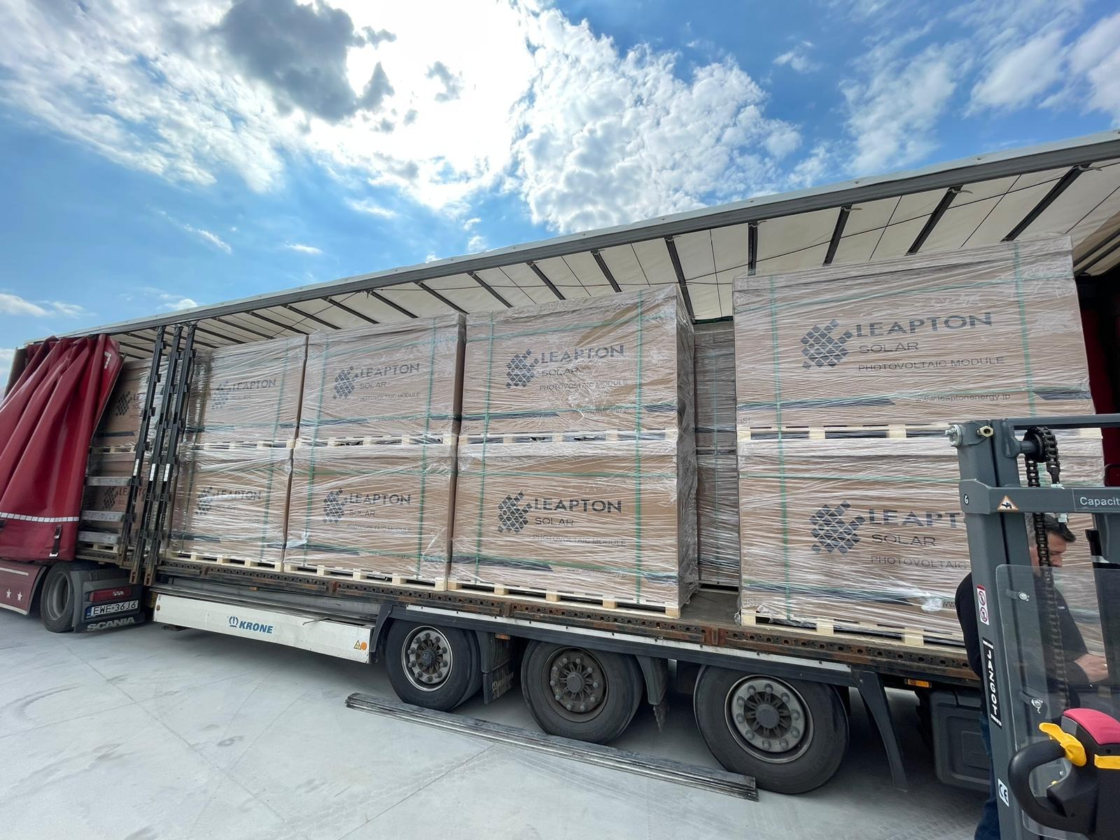 Ciężarówka z naczepą załadowana paletami z modułami fotowoltaicznymi Leapton Solar, zabezpieczonymi folią stretch, na tle błękitnego nieba z chmurami. Widoczny wózek widłowy przygotowujący się...