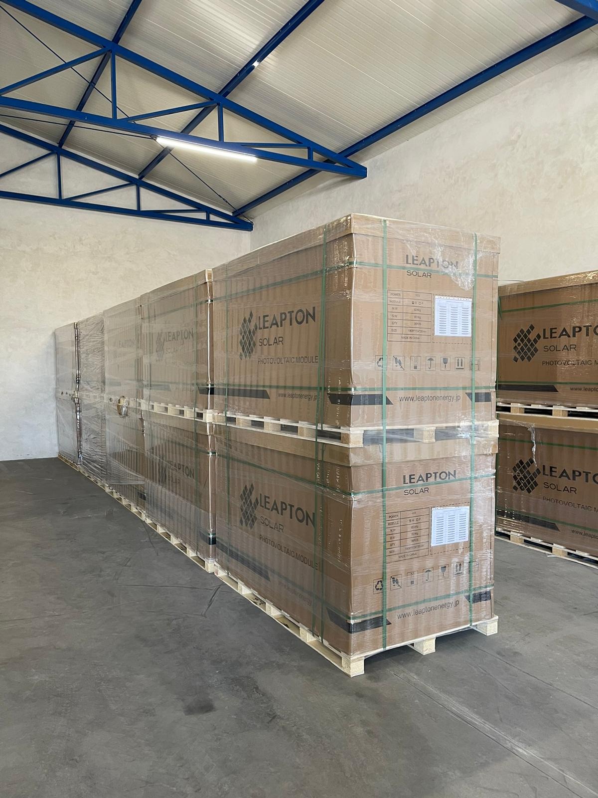 Palety z zapakowanymi modułami fotowoltaicznymi Leapton Solar, zabezpieczone folią, składowane w magazynie z metalową konstrukcją dachu.