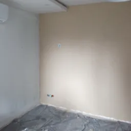 Pokój w trakcie prac przygotowawczych: ściany częściowo pokryte gładzią, podłoga zabezpieczona folią malarską, widoczne gniazdka elektryczne.