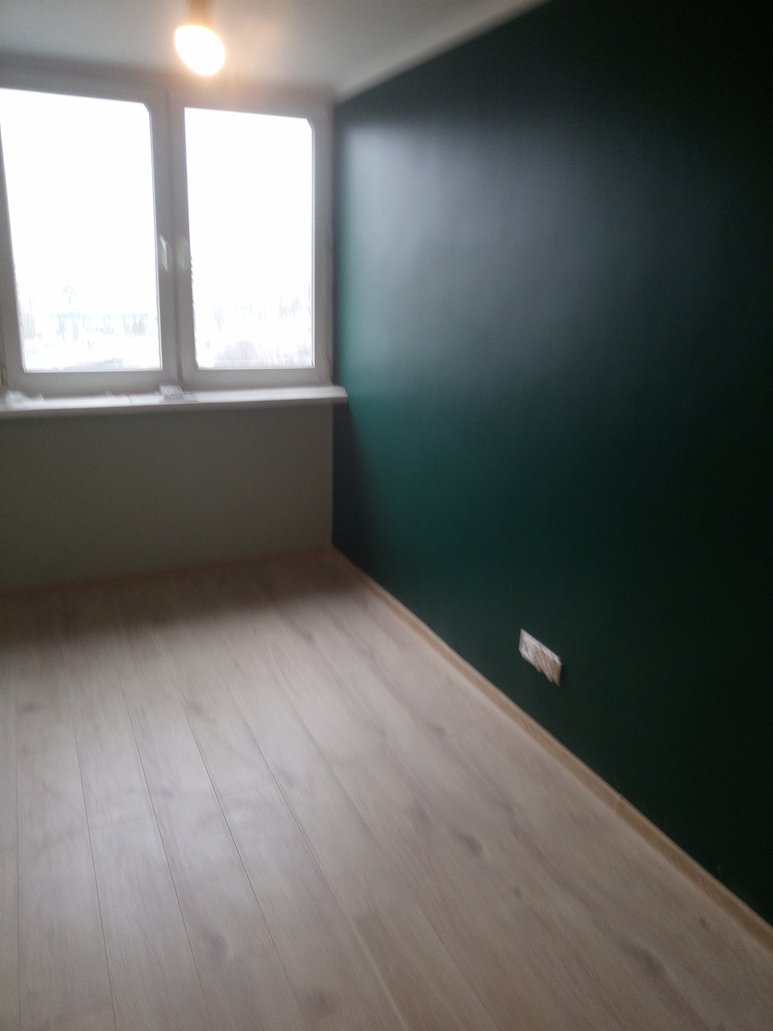 Pomalowane na ciemnozielono wnętrze pokoju z jasną podłogą i oknem; widoczny fragment ściany pod oknem w jaśniejszym odcieniu zieleni oraz gniazdko elektryczne.