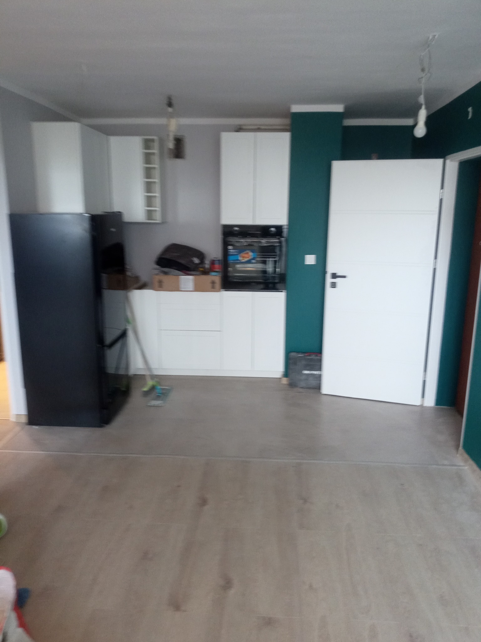 Wnętrze kuchni w trakcie prac wykończeniowych, widoczne białe szafki kuchenne, czarna lodówka, mop, kartony na blacie, ściana pomalowana na ciemny zielony kolor, białe drzwi i podłoga z paneli.