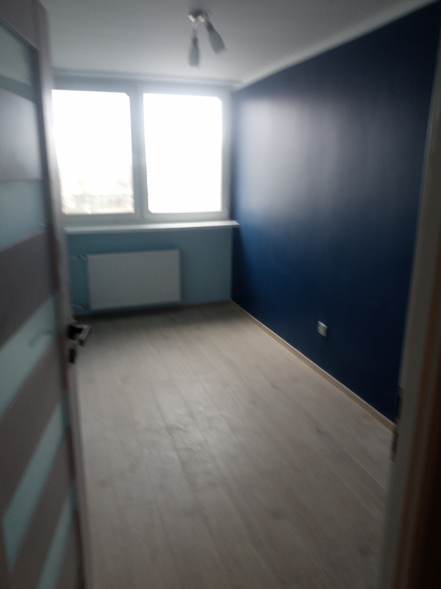 Pomalowane na granatowo wnętrze pokoju z jasnymi panelami podłogowymi, widok zza uchylonych drzwi z oknem i grzejnikiem w tle.