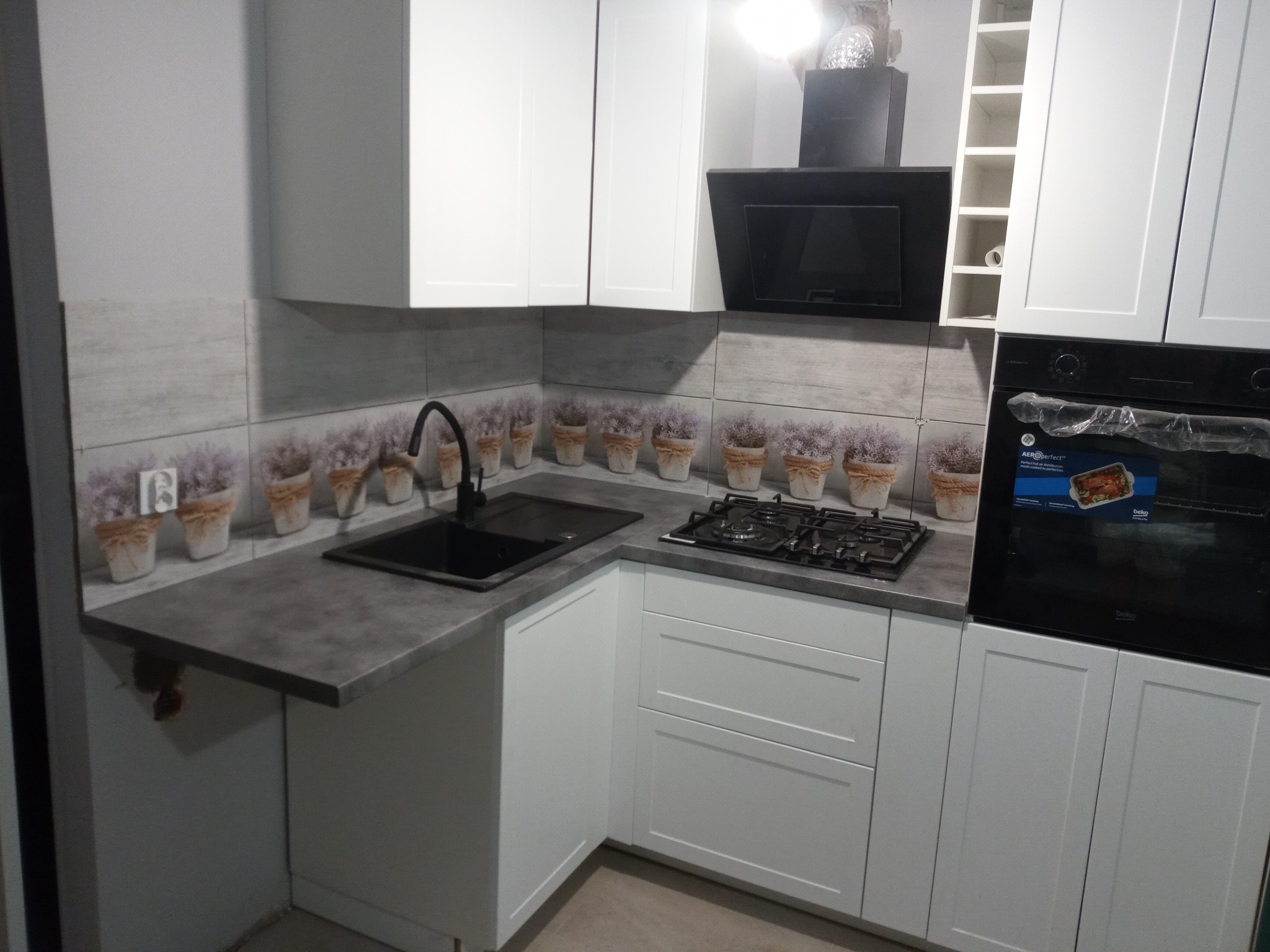 Nowoczesna kuchnia z białymi szafkami, szarym blatem i płytkami z motywem lawendy w doniczkach, czarnym zlewem i płytą gazową.