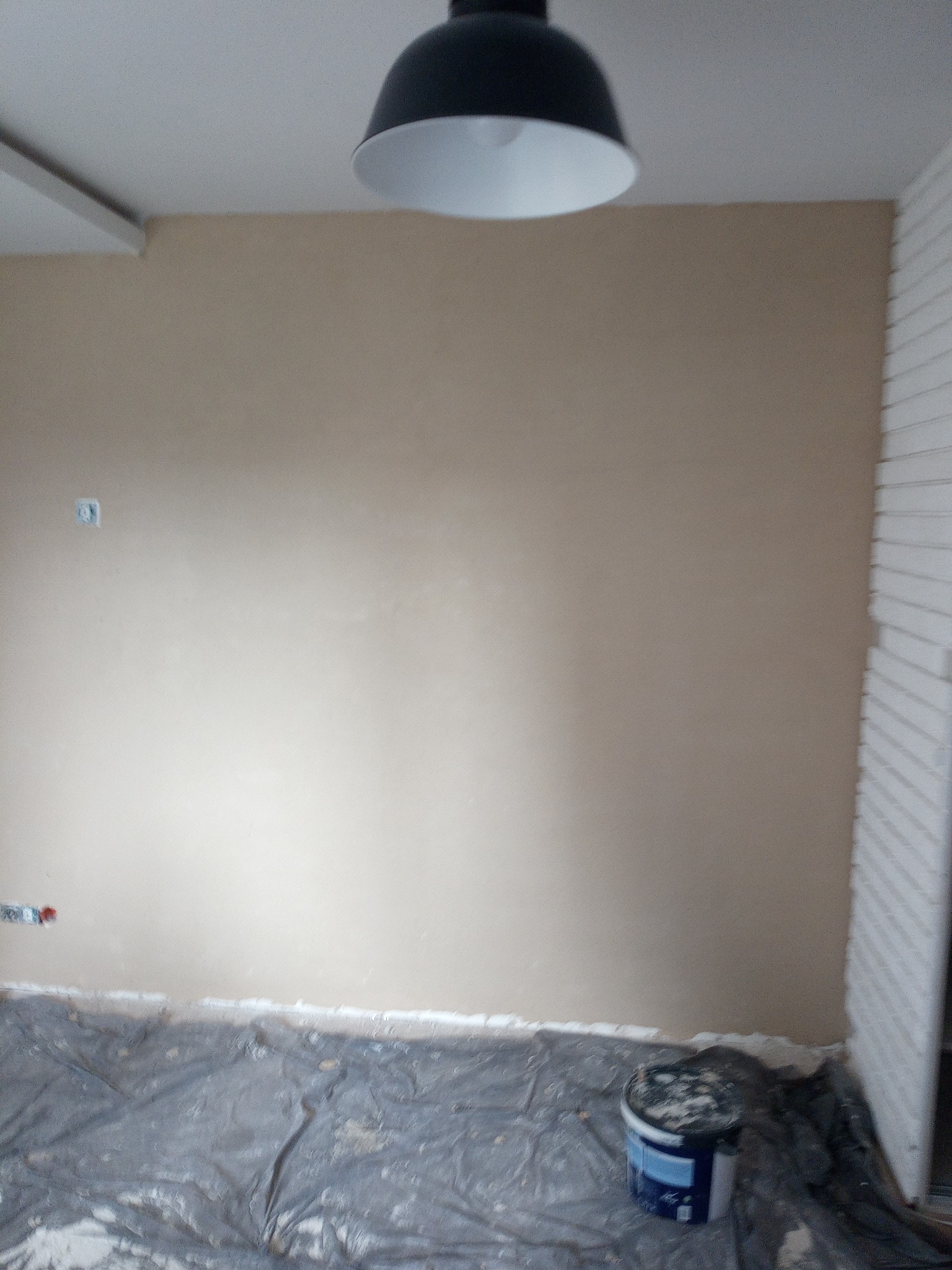 Ściana po szpachlowaniu w beżowym odcieniu, zabezpieczona folią podłoga, widoczne puszki elektryczne i wiadro z resztkami szpachli, lampa sufitowa w tle.