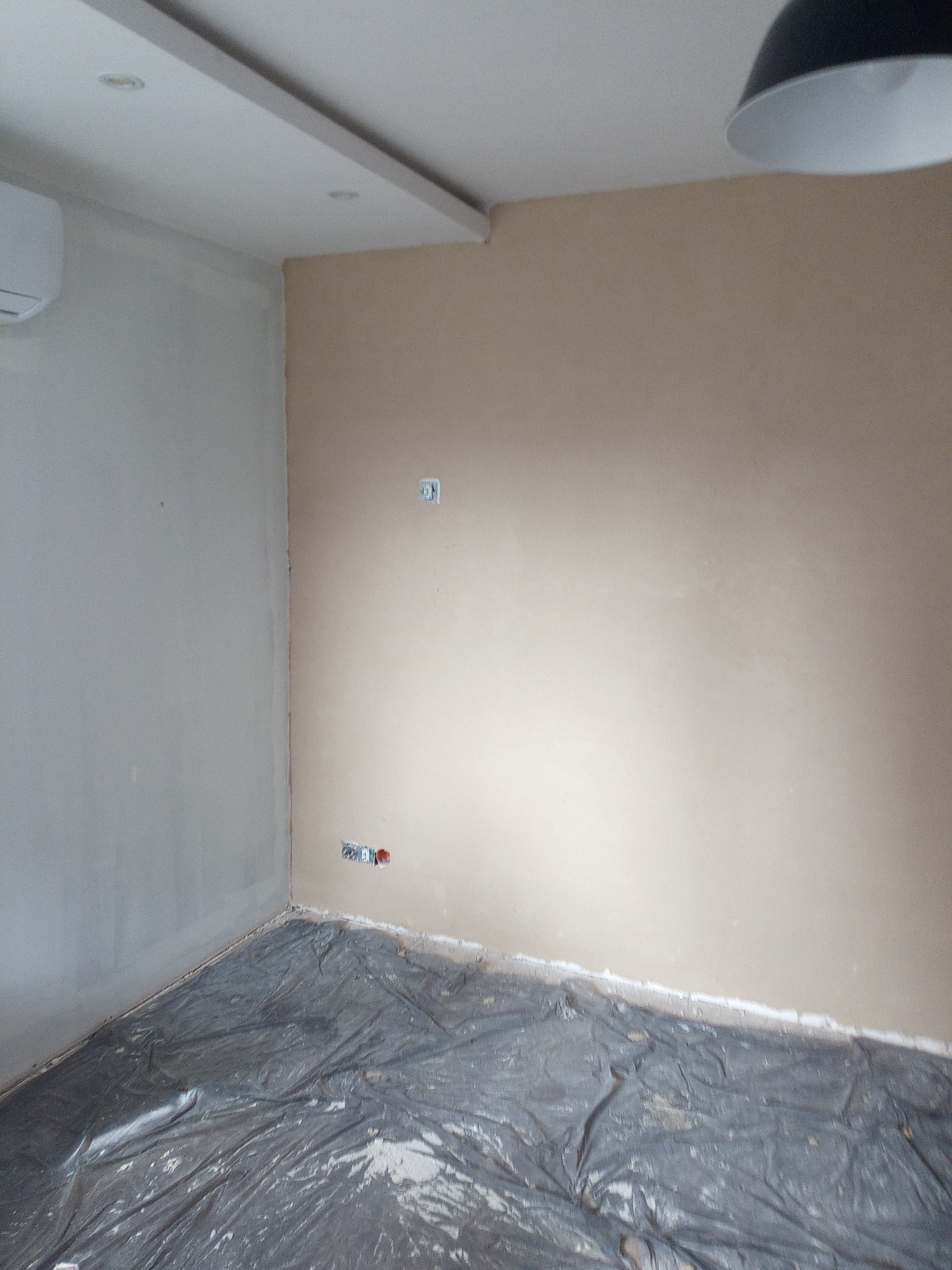 Pokój w trakcie prac przygotowawczych: ściany częściowo pokryte gładzią, podłoga zabezpieczona folią malarską, widoczne gniazdka elektryczne.