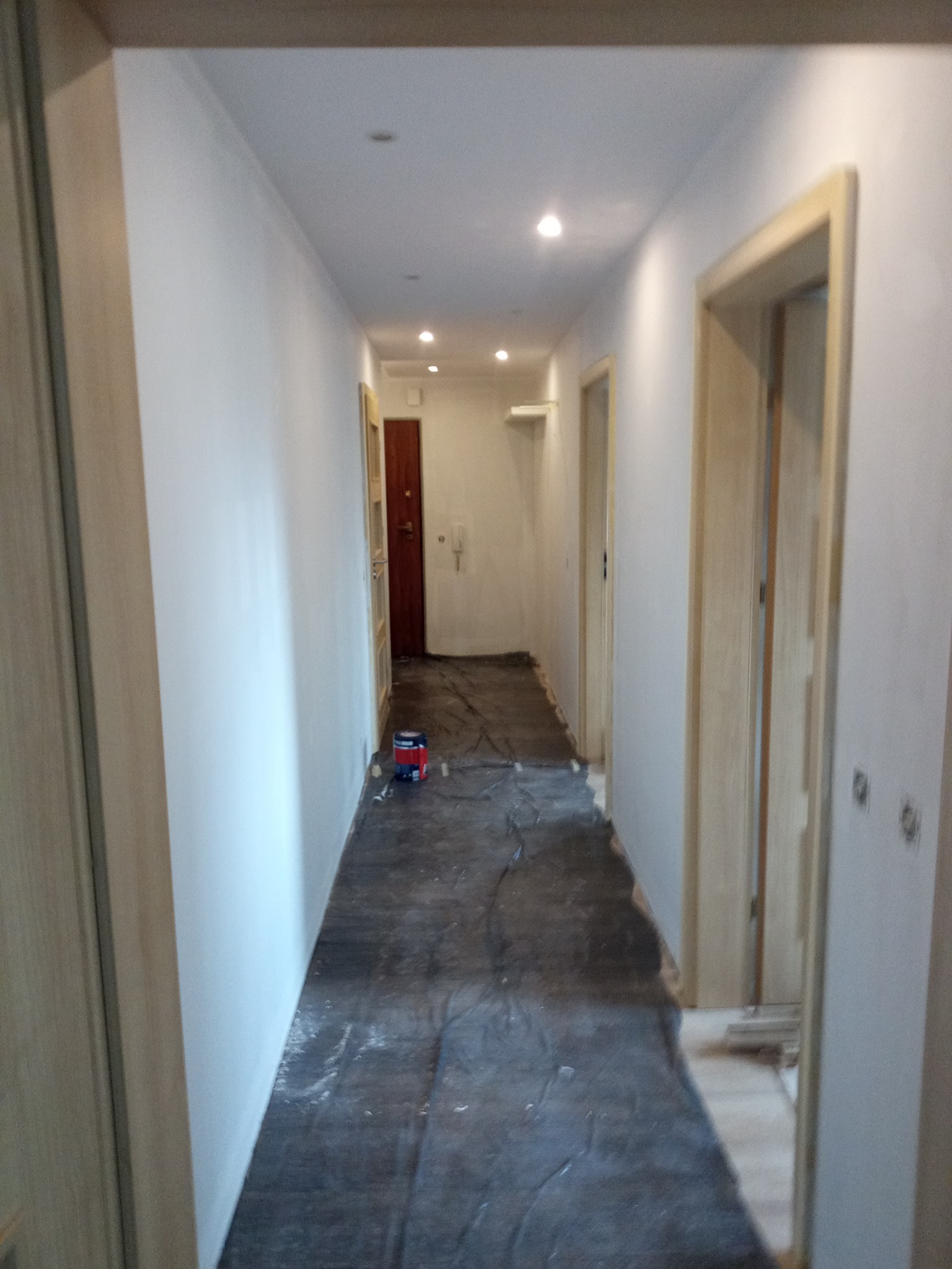 Korytarz w trakcie prac wykończeniowych, ściany po malowaniu na biało, podłoga zabezpieczona folią, widoczna puszka z farbą i wałek malarski.
