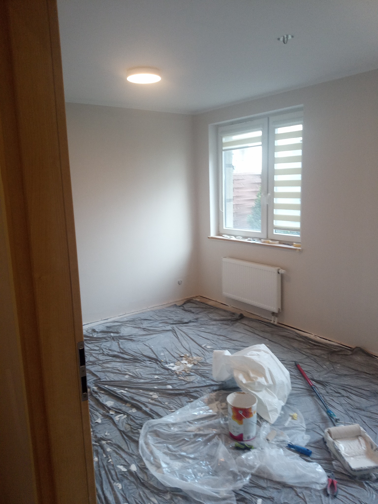 Pokój w trakcie przygotowań do malowania: zabezpieczona podłoga folią, widoczne narzędzia malarskie, pędzel, kuweta z farbą, wiadro z motywem kwiatowym.