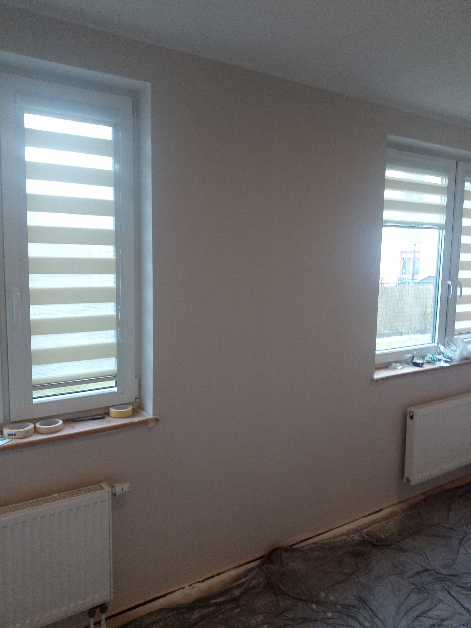 Świeżo pomalowane ściany w pokoju z dwoma oknami z żaluzjami, zabezpieczona folią podłoga, grzejniki zamontowane pod oknami, widoczne ślady taśmy malarskiej na parapetach.