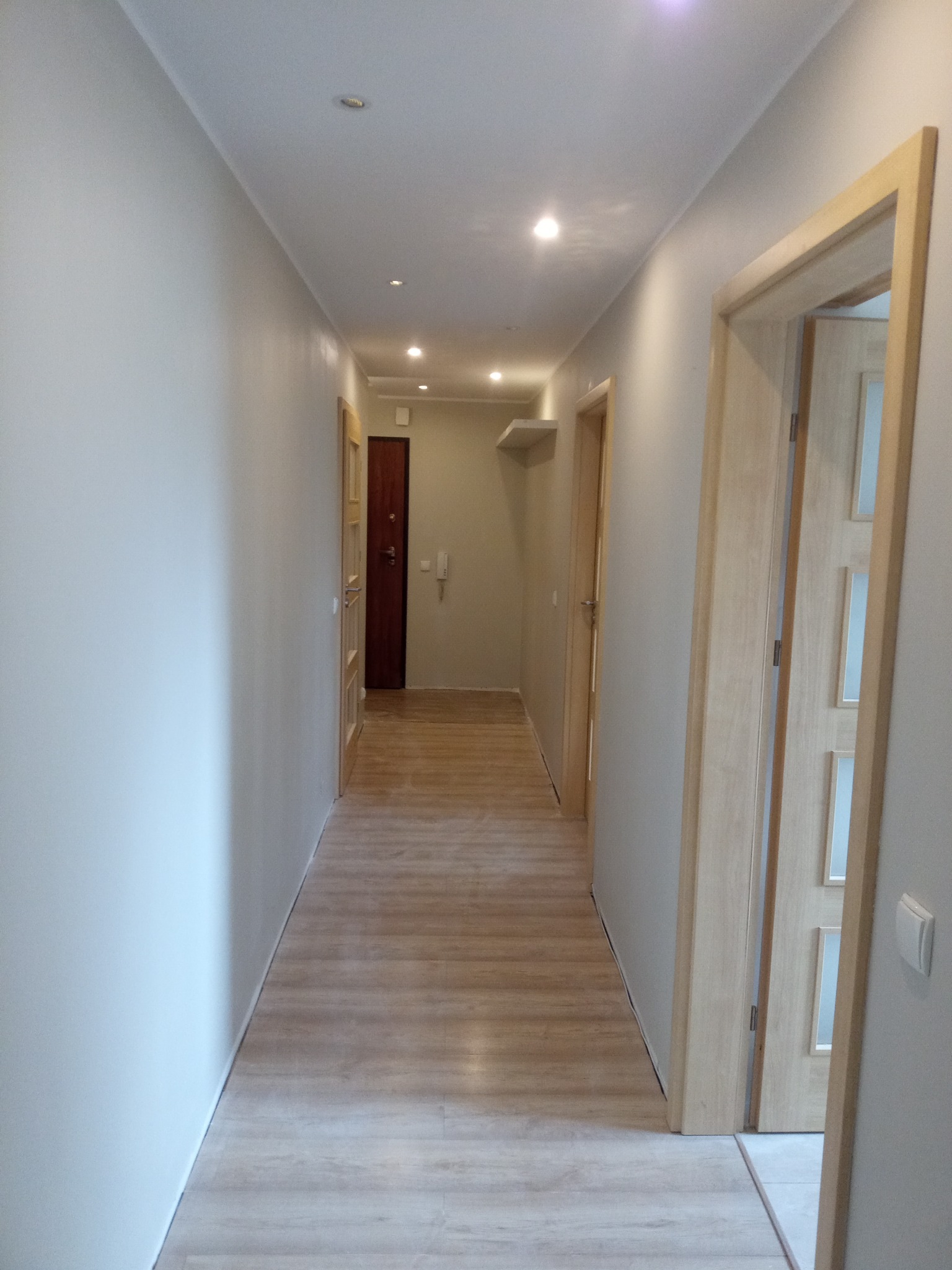 Wyremontowany korytarz z panelami podłogowymi w odcieniach drewna, świeżo pomalowanymi ścianami w kolorze jasnoszarym i drewnianymi drzwiami z przeszkleniami.