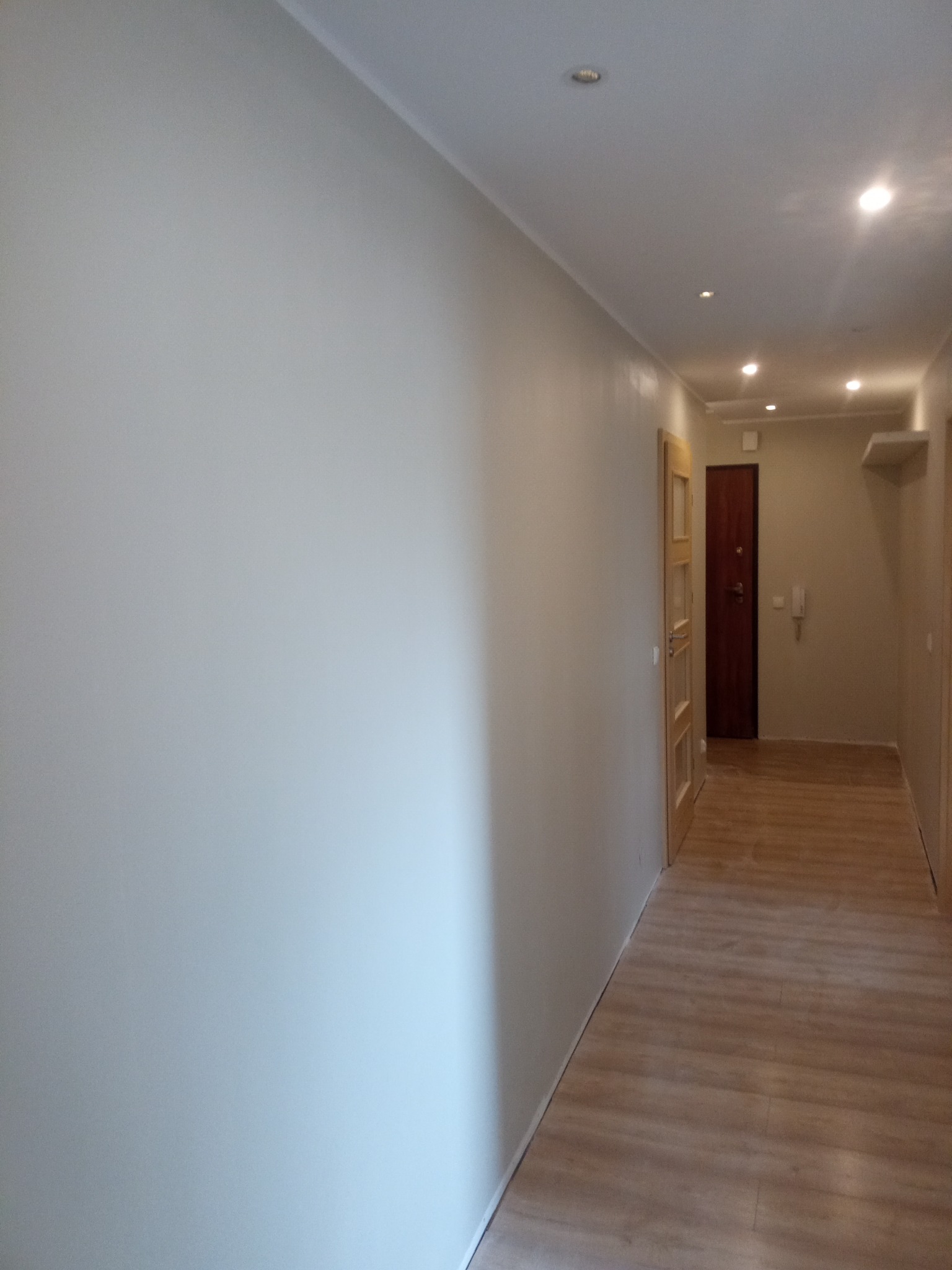 Jasne, świeżo pomalowane ściany w korytarzu z drewnianą podłogą, widoczne drzwi i oświetlenie punktowe w suficie.