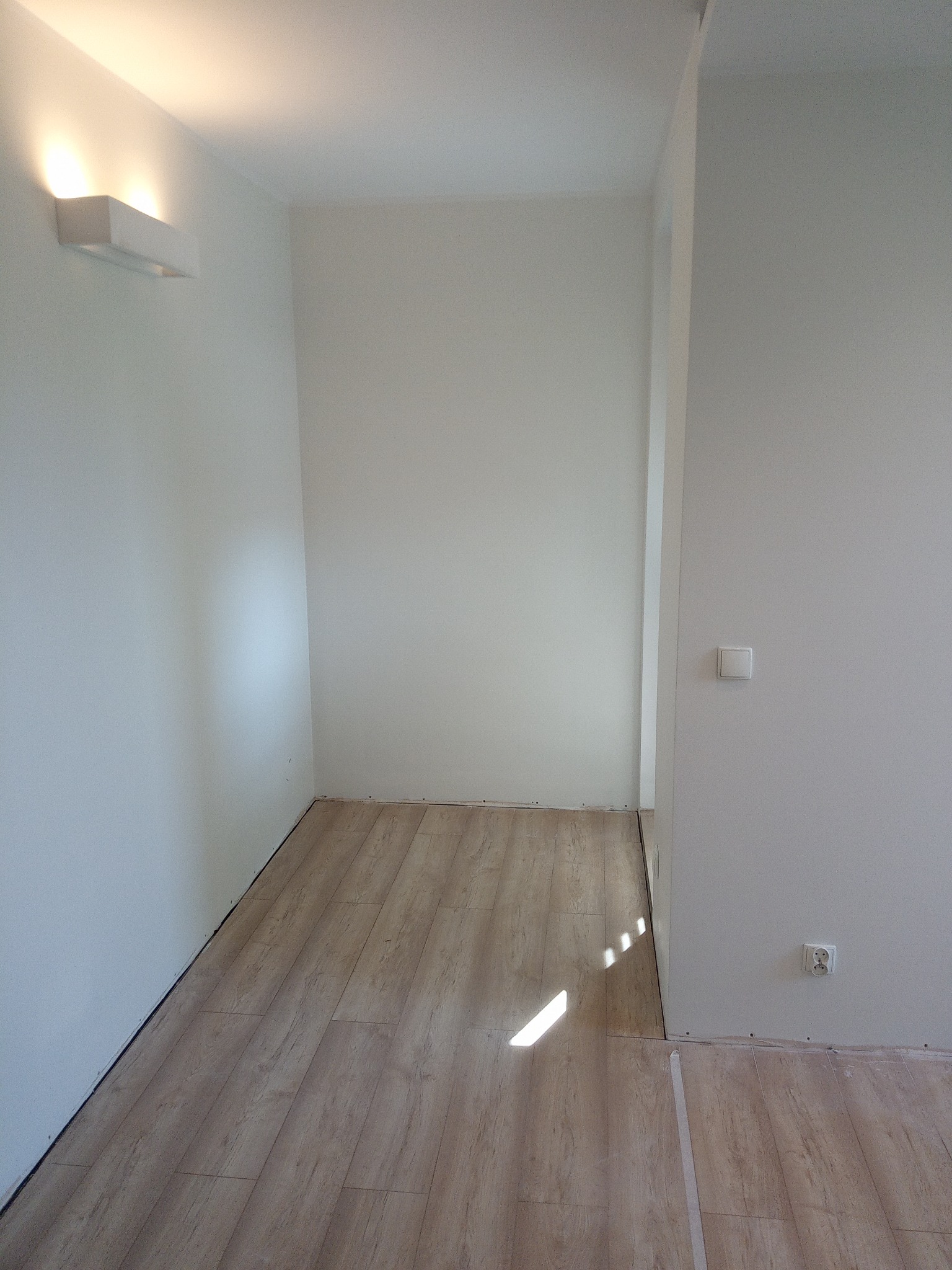 Fragment pokoju z ułożoną podłogą z paneli imitujących drewno, białe ściany z zamontowanym oświetleniem ściennym i gniazdkiem elektrycznym, widoczne ślady po pracach wykończeniowych.
