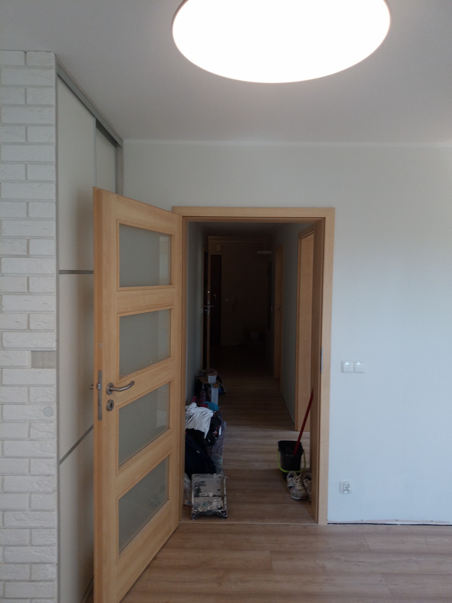 Otwarta drewniana drzwi z przeszkleniami, widok na korytarz z elementami wyposażenia i przygotowań do prac, jasne pomieszczenie z białymi ścianami i podłogą imitującą drewno.