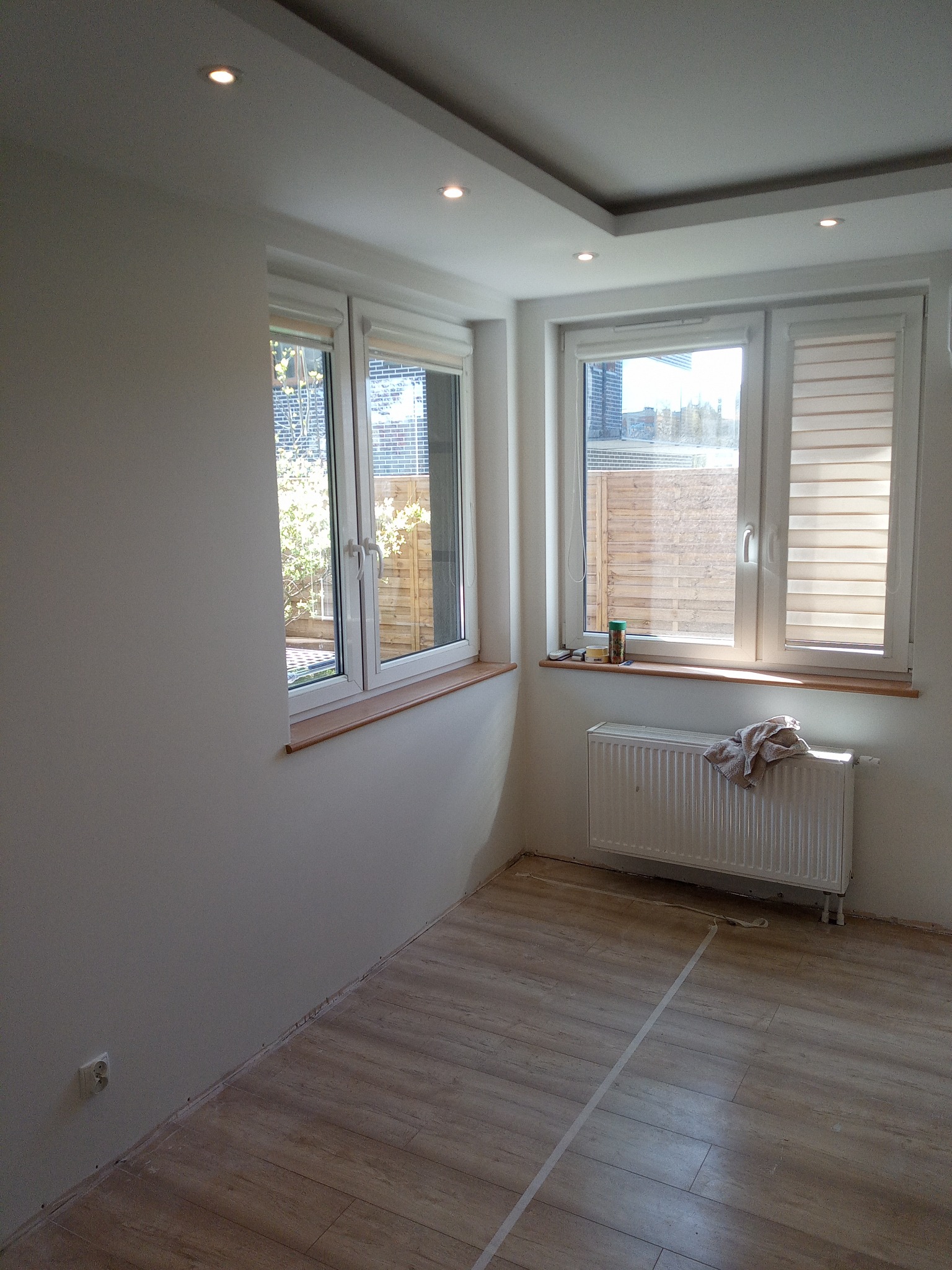 Wnętrze pokoju we Wrocławiu po świeżym malowaniu ścian na biało, z drewnopodobną podłogą, białym grzejnikiem z ręcznikiem, dwoma oknami z roletami i widocznymi śladami taśmy malarskiej na podłodze.