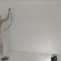 Pracownik w kombinezonie ochronnym maluje ścianę w pomieszczeniu za pomocą pistoletu natryskowego; podłoga zabezpieczona folią, widoczne gniazdka elektryczne.