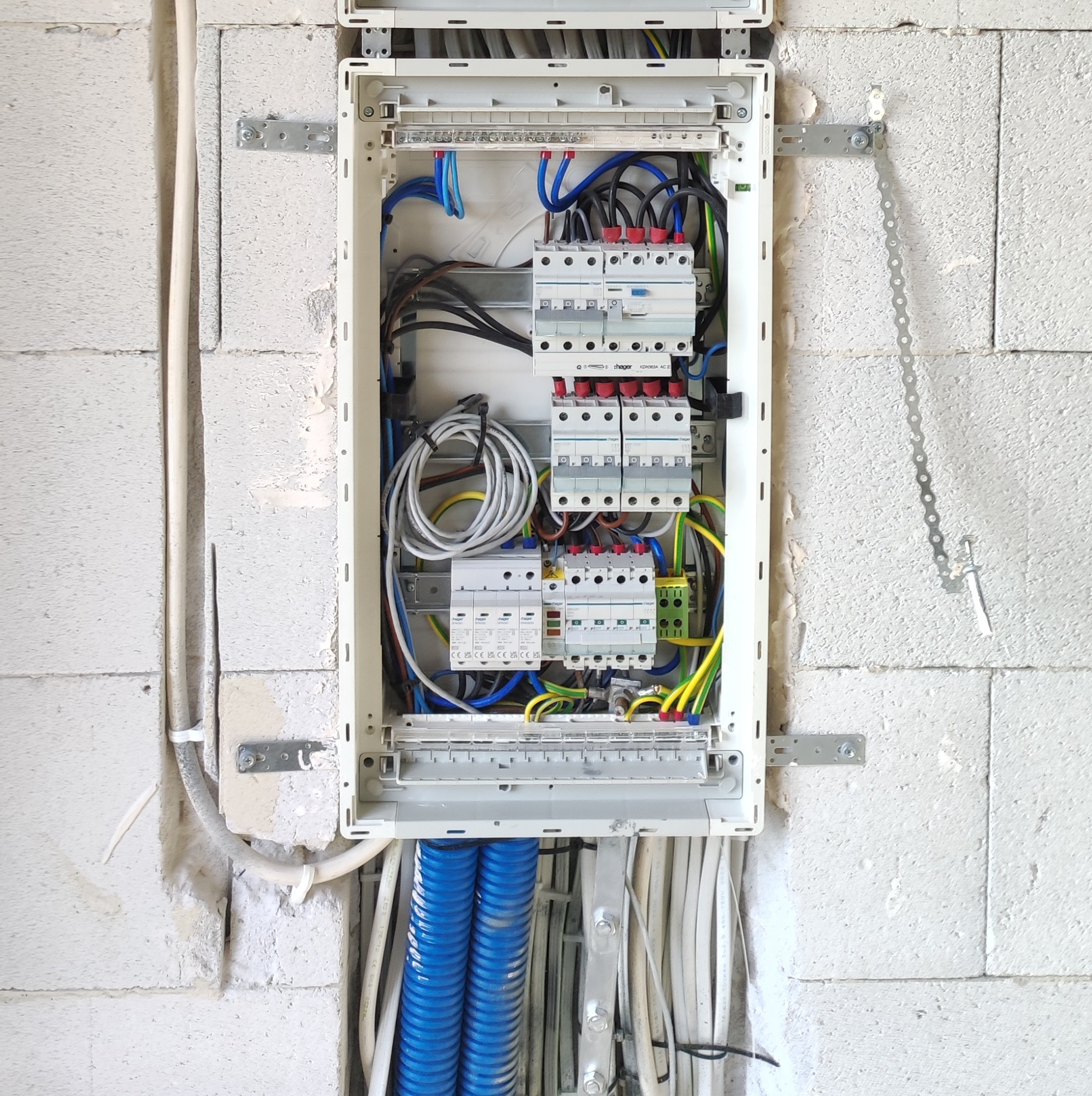Rozdzielnica elektryczna w stanie surowym, widoczne kable, bezpieczniki i przewody w korytkach, zamontowana na ścianie z białych bloczków.