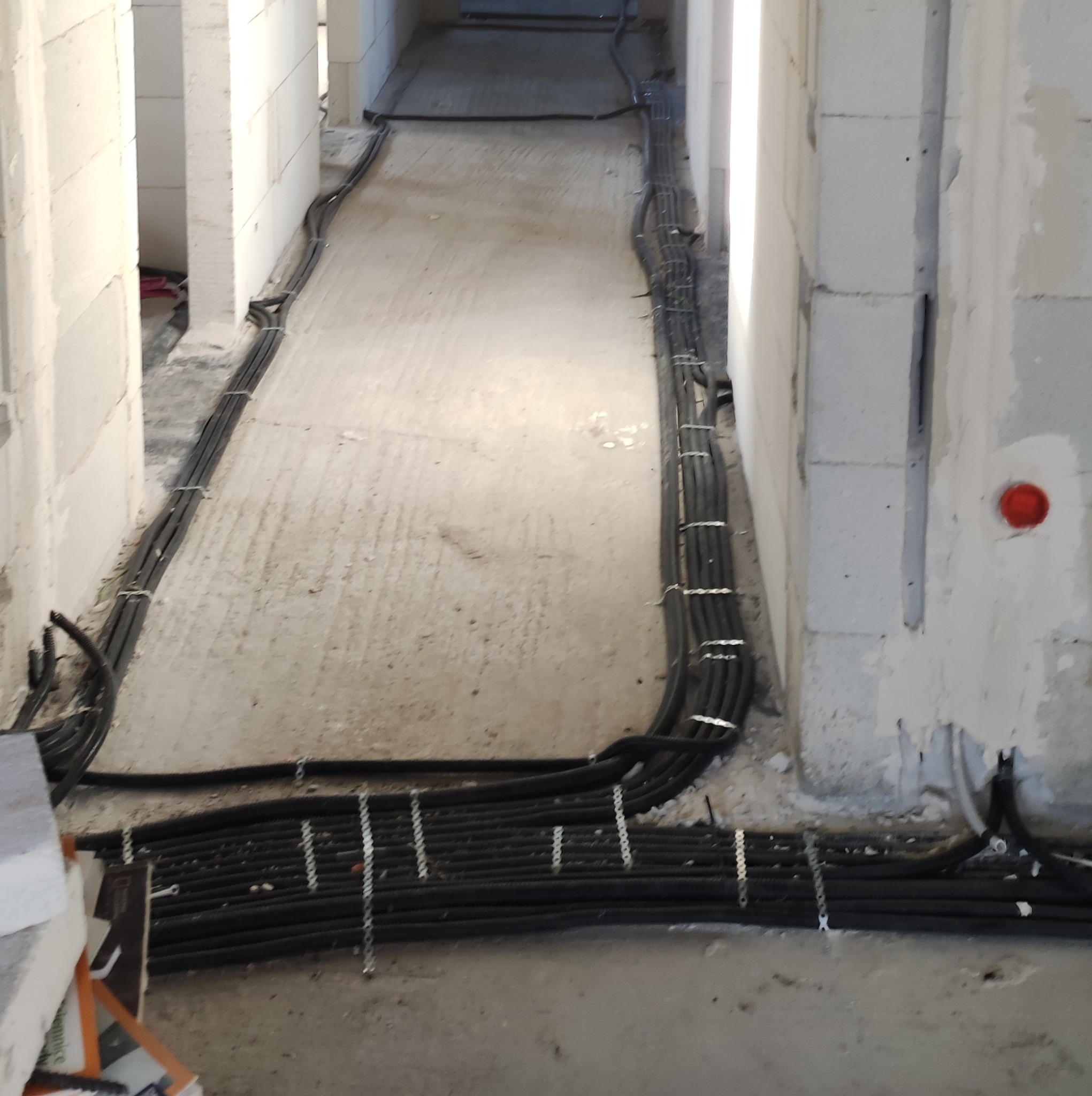 Kable elektryczne w peszlach ułożone wzdłuż ścian w surowym stanie budynku z białych bloczków, przymocowane metalowymi uchwytami do podłoża.