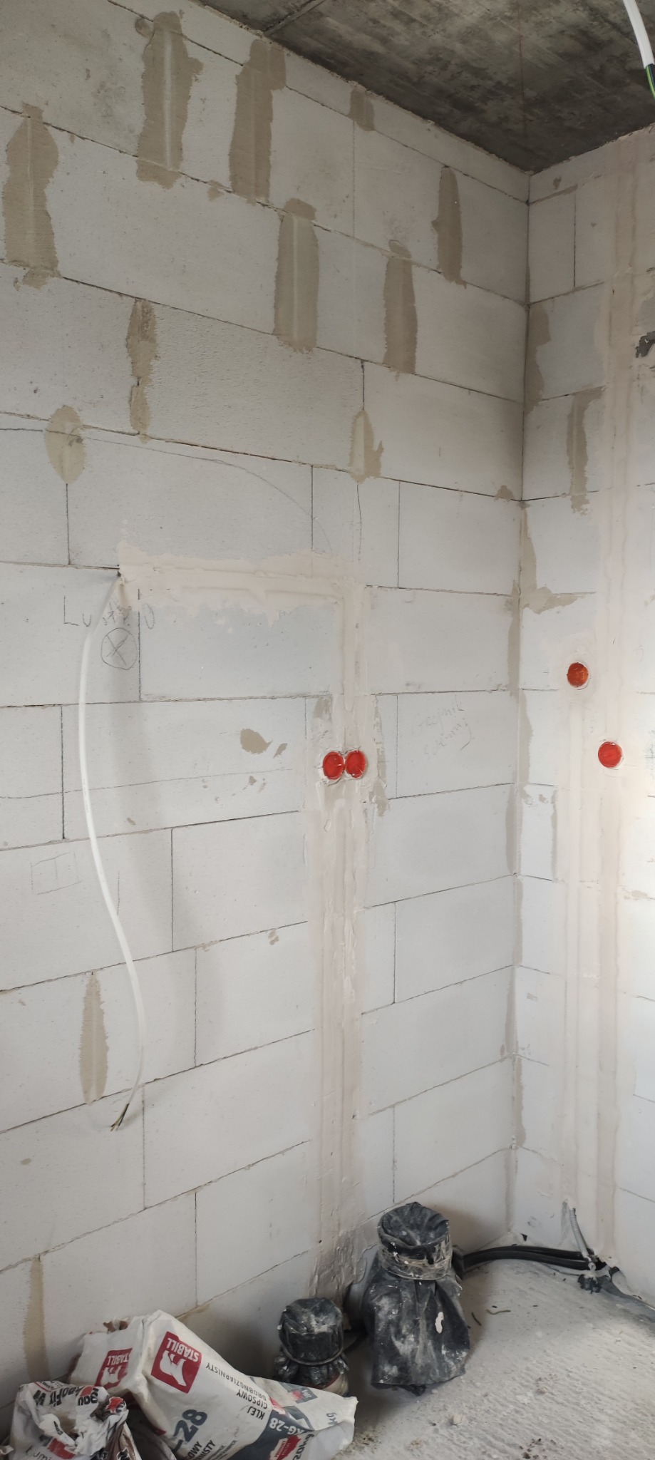 Ściana z betonu komórkowego z widocznymi przewodami elektrycznymi w peszlach, zamurowanymi puszkami elektrycznymi i śladami po zaprawie murarskiej oraz workami z klejem na podłodze.