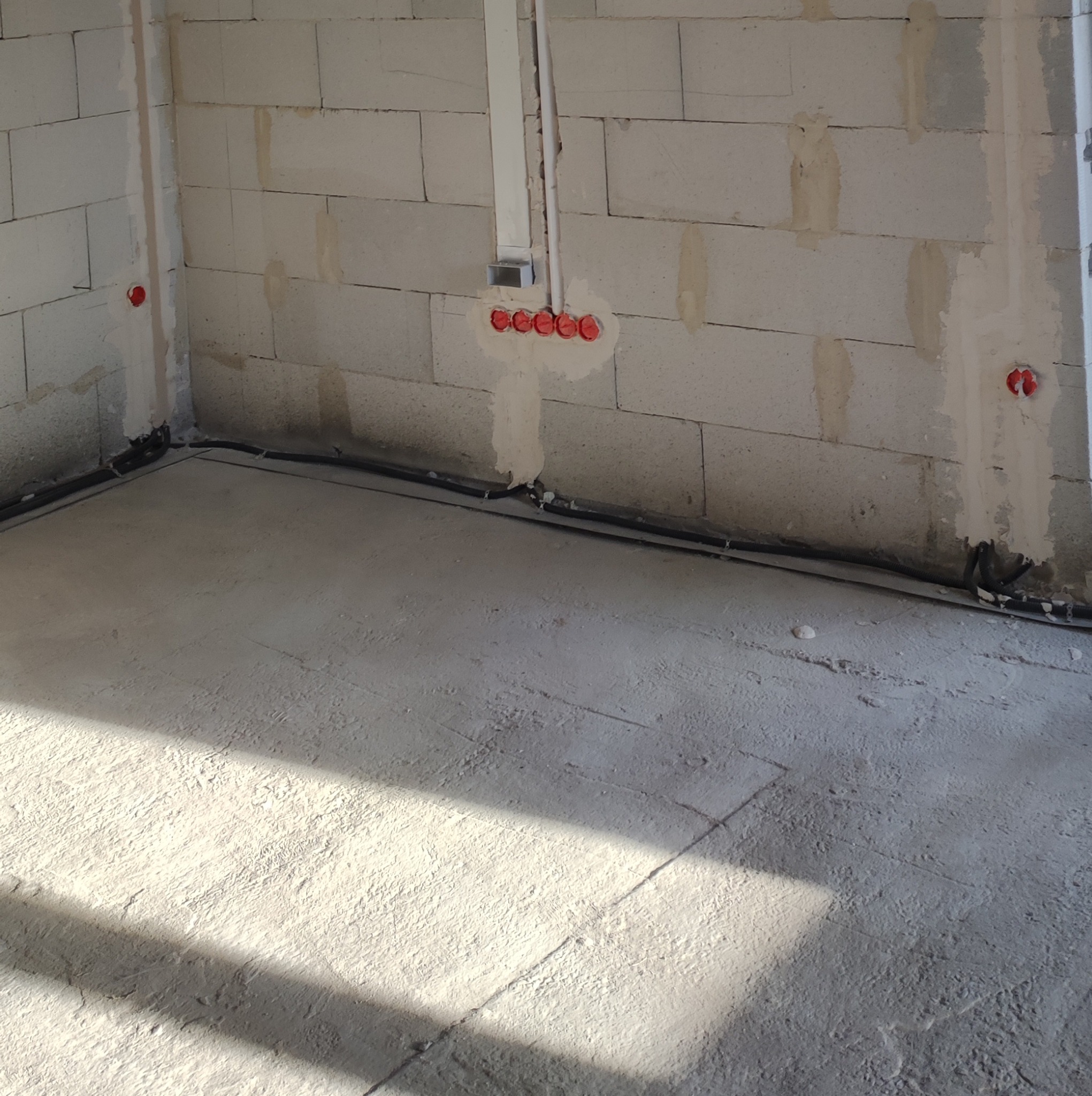 Surowy stan wnętrza z widoczną instalacją elektryczną w ścianach z betonu komórkowego, czarnymi przewodami ułożonymi wzdłuż podłogi i wstępnie przygotowaną wylewką podłogową w słonecznym oświetleniu.