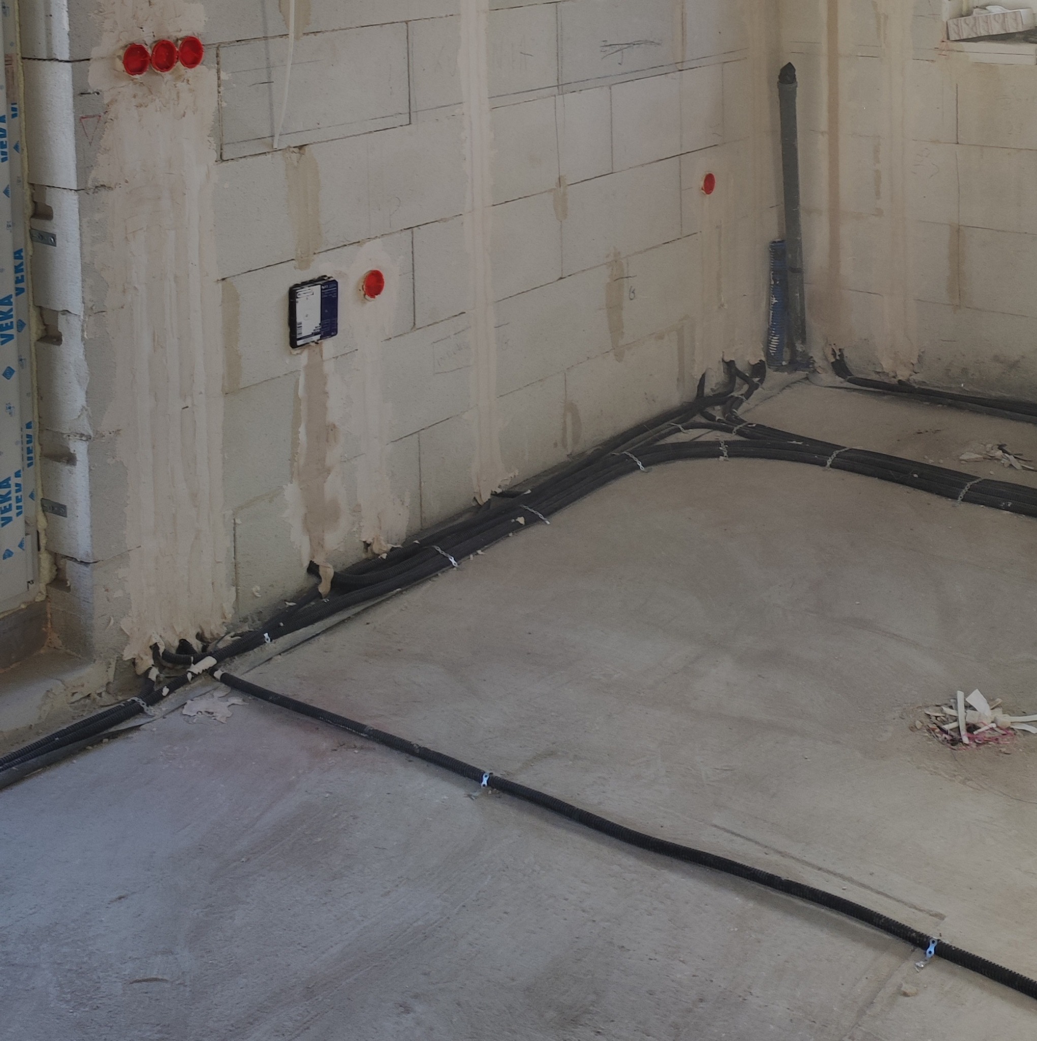 Surowy stan wnętrza z widocznymi przewodami elektrycznymi w peszlach prowadzonymi po podłodze i wzdłuż ścian z betonu komórkowego, czerwone puszki instalacyjne w ścianie, pion kanalizacyjny w rogu...