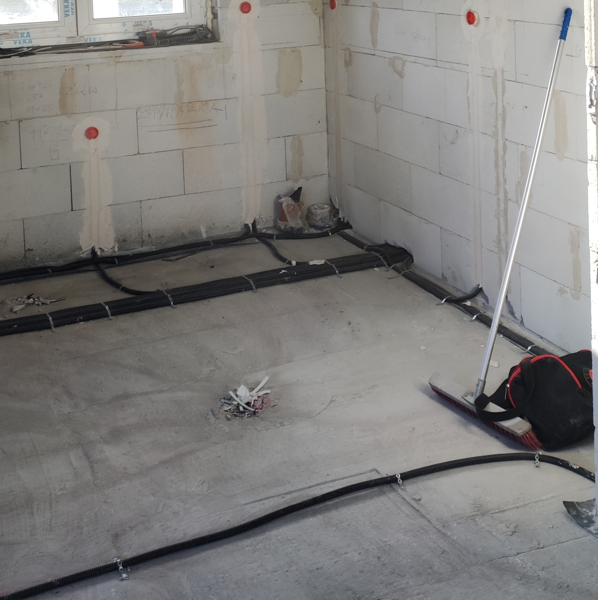 Surowy stan wnętrza z instalacją elektryczną w peszlach mocowanych do podłoża, widoczne ślady po kuciu w ścianach z betonu komórkowego, czerwone puszki elektryczne w ścianach, szczotka...