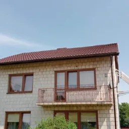 Dwupiętrowy dom z jasnej cegły z nowym, brązowo-czerwonym dachem z dachówki, brązowymi ramami okiennymi i balkonem z metalową balustradą.